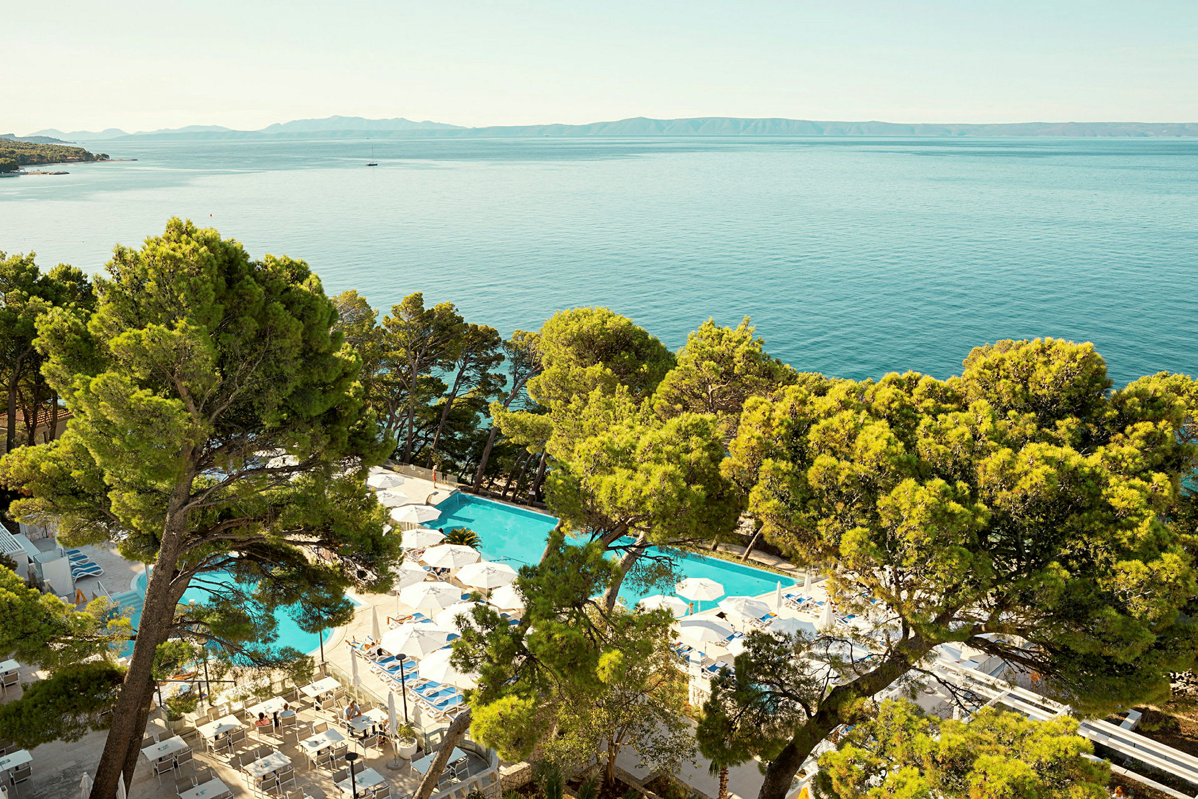 Utsikt över en ljusblå pool, omgiven av parasoller och solsängar, med gröna träd och havet i bakgrunden. (AI-genererad text)