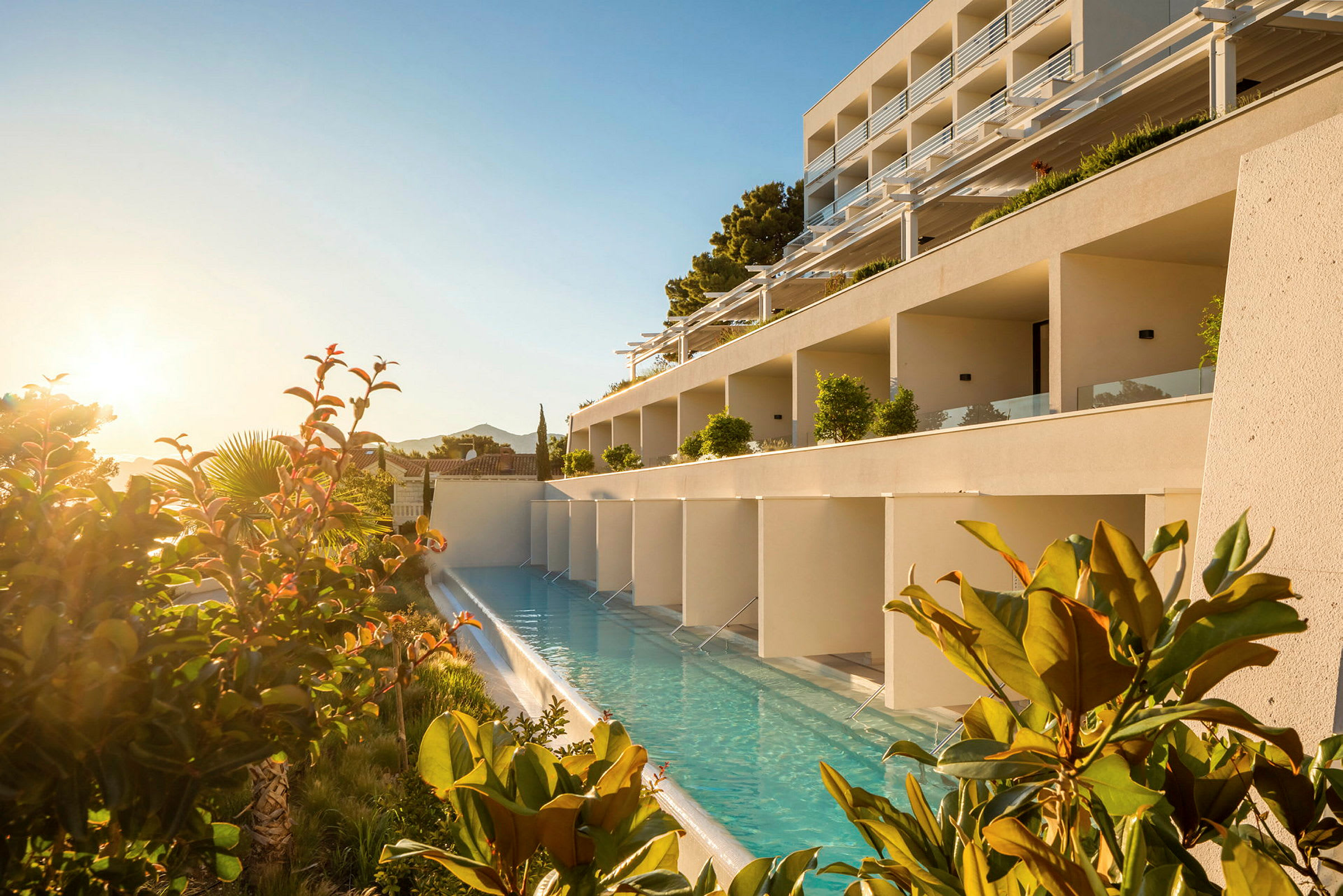 En hotellbyggnad med en längsgående pool framför, omgiven av växter och soluppgång. (AI-genererad text)