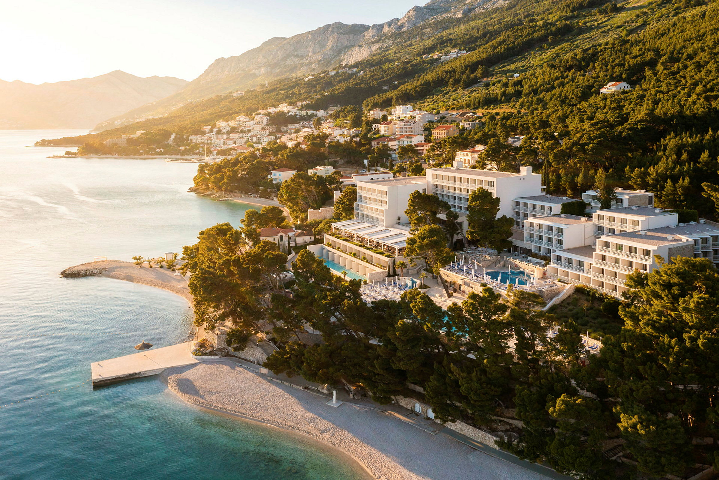 Luftfoto av hotell vid kusten och strand, omgiven av träd och med berg i bakgrunden. (AI-genererad text)