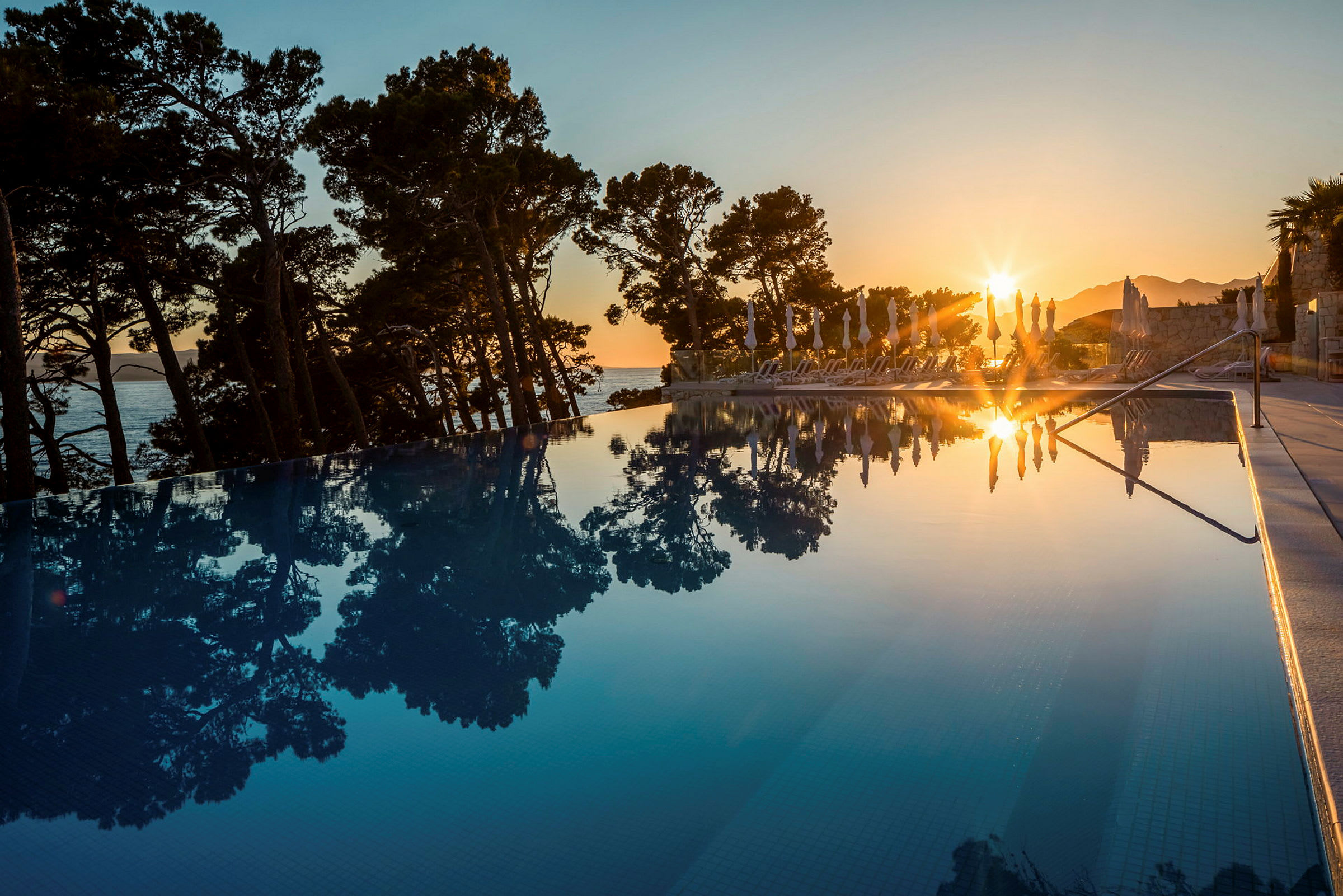 En solnedgång reflekteras i en pool, omgiven av tallar och solstolar. (AI-genererad text)