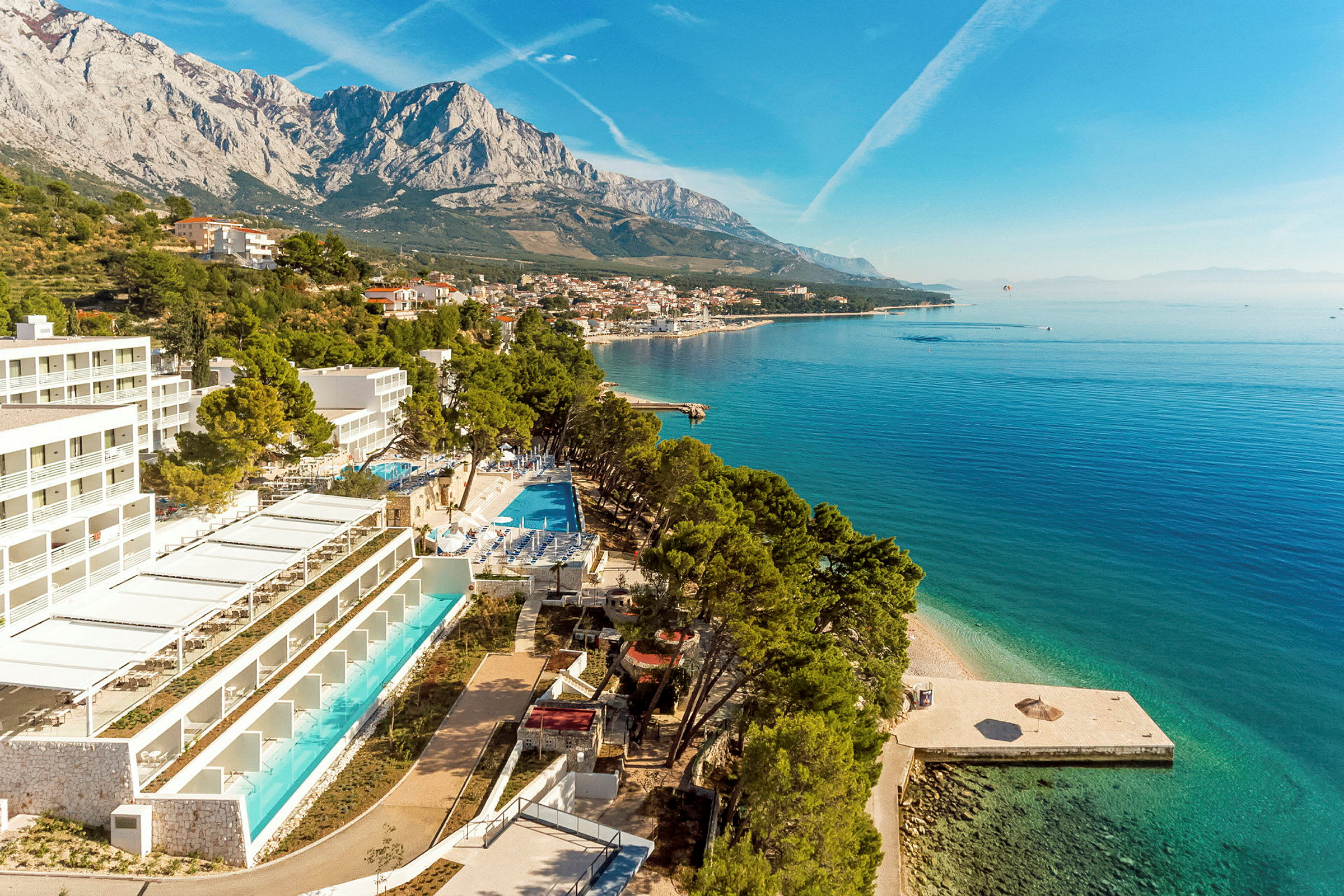 Vy över kustnära hotell med pool nära havet och berg i bakgrunden. (AI-genererad text)