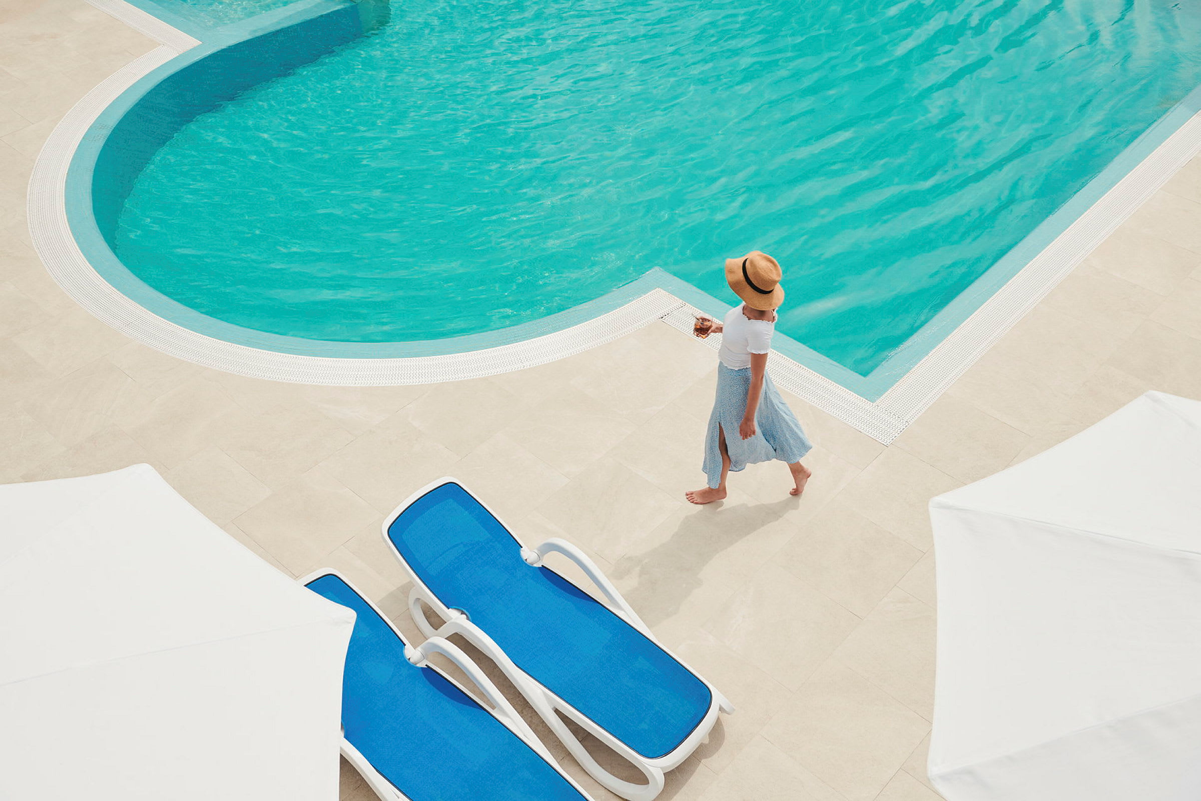Kvinna promenerar nära en pool med blå solstolar och vita parasoller. (AI-genererad text)