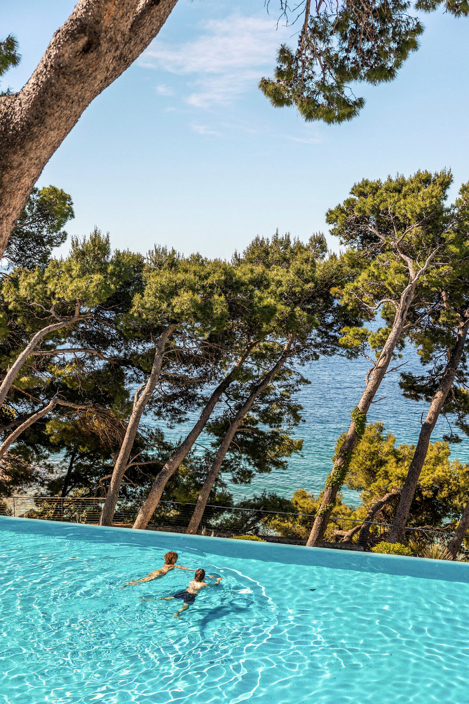 Två personer simmar i en pool med utsikt över havet och trädkronor. (AI-genererad text)