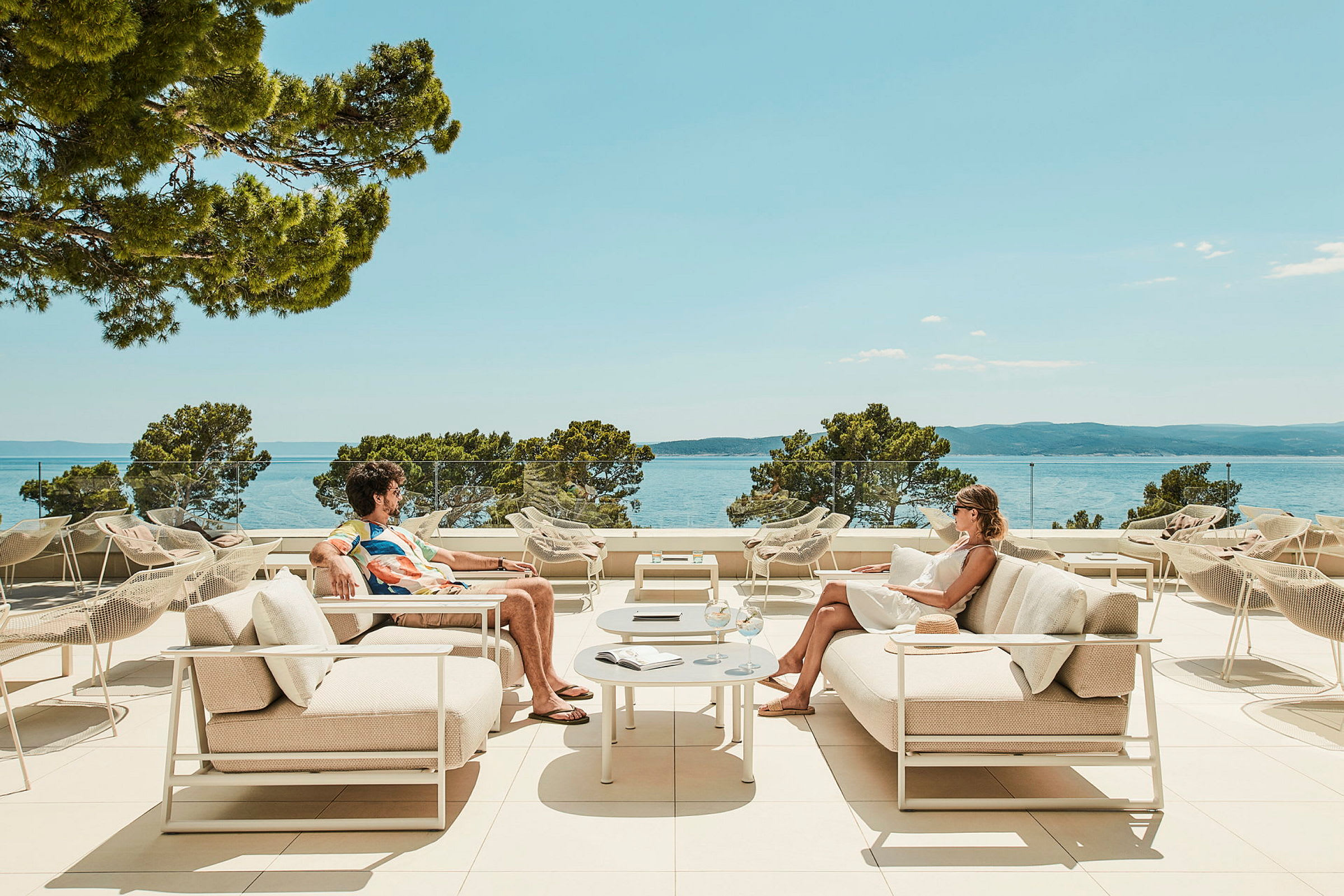Två personer sitter på moderna uteplatsmöbler med utsikt över havet i bakgrunden. (AI-genererad text)