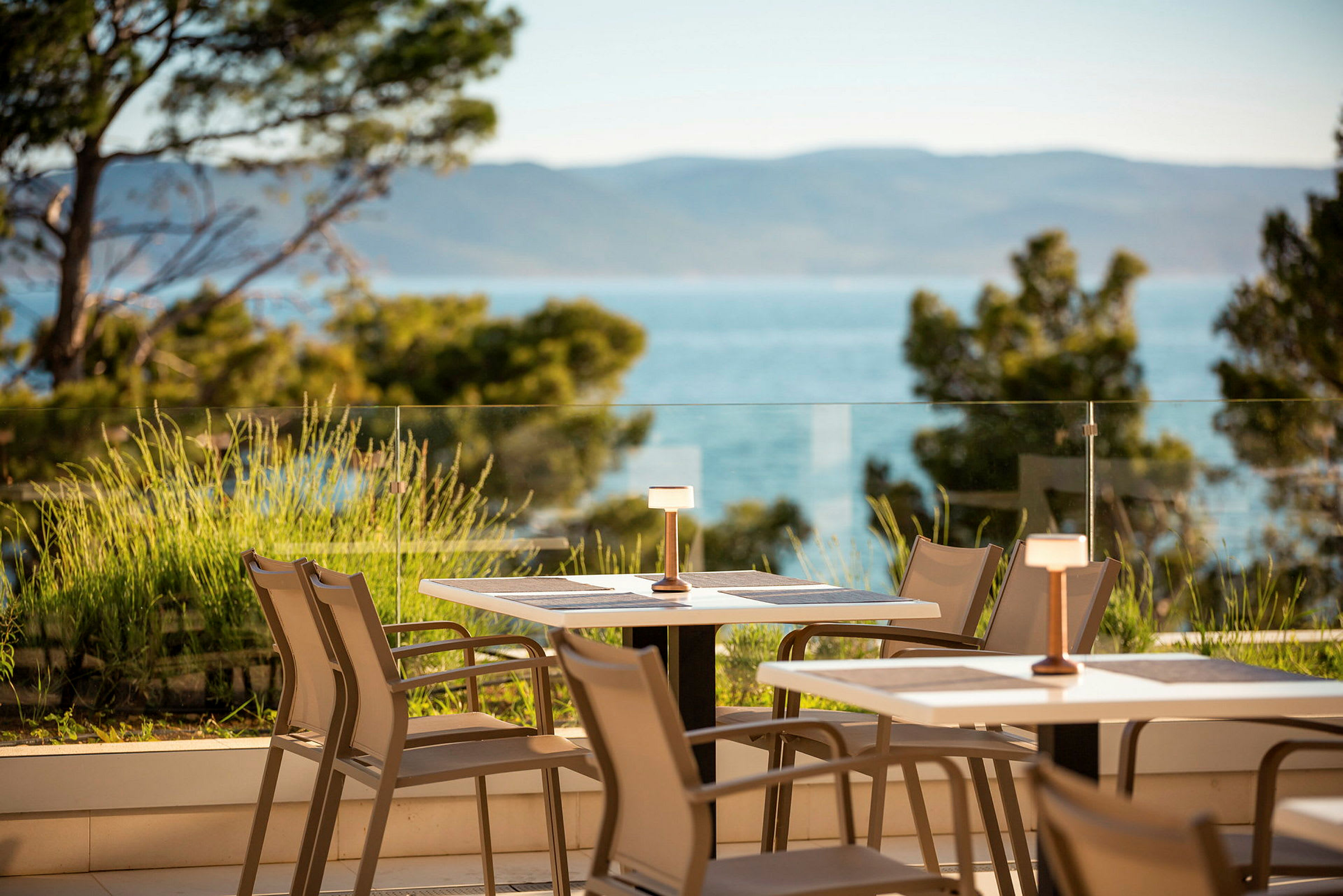 Utsikt över en terrass med bord och stolar, omgivet av grönska och med havet i bakgrunden. (AI-genererad text)