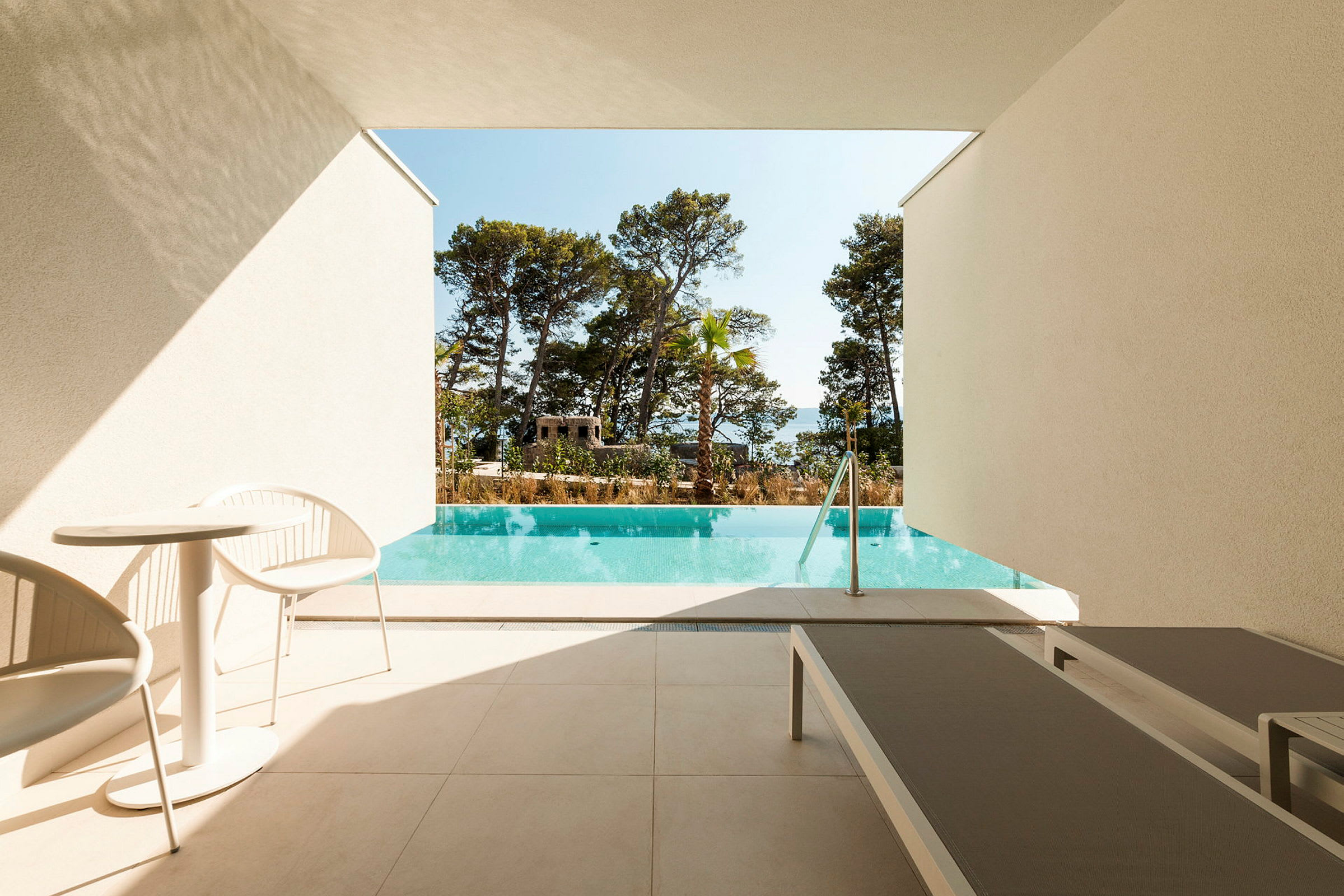 Bild av en terrass med en liten pool i förgrunden, två solstolar och ett möbelset med två stolar och ett bord. Träd och klar himmel syns i bakgrunden. (AI-genererad text)