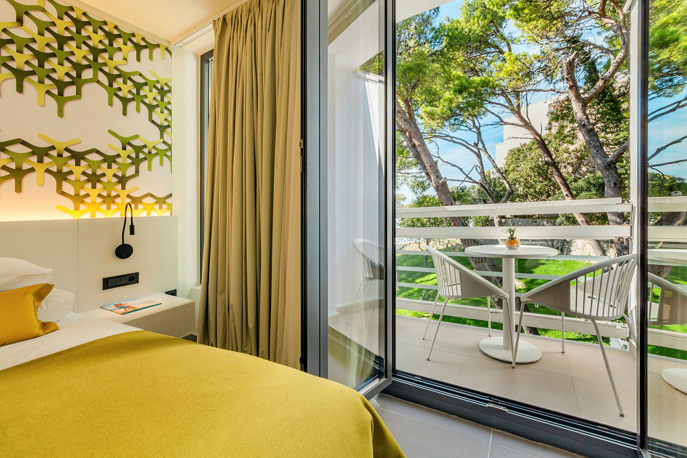 Interiören visar ett modernt sovrum med gul inredning och en balkon med stolar och ett bord. Utsikt över grönskande träd utanför. (AI-genererad text)