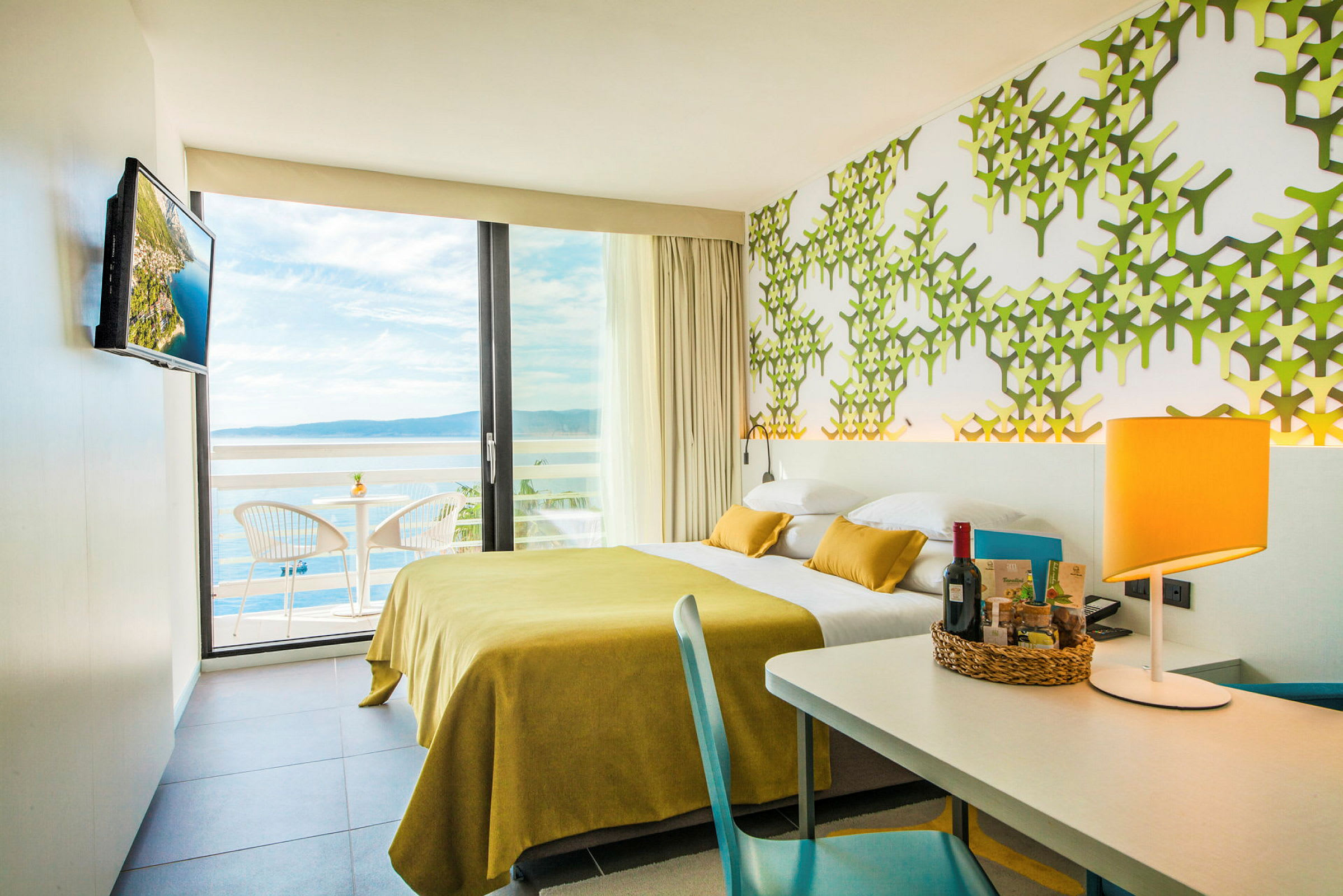 Hotellrum med modern inredning, gul sängöverkast, grön mönstrad vägg och utsikt över havet från balkongen. (AI-genererad text)