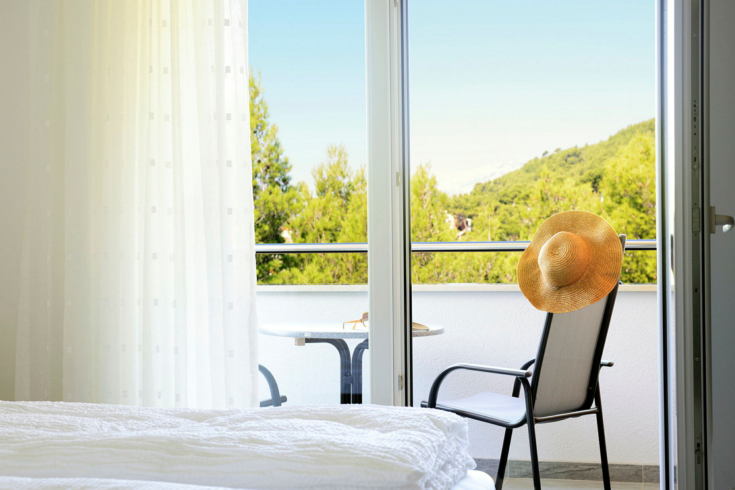 Ett hotellrum med utsikt över en balkong där en stol med en hatt och ett bord står, samt grönskande träd i bakgrunden. (AI-genererad text)