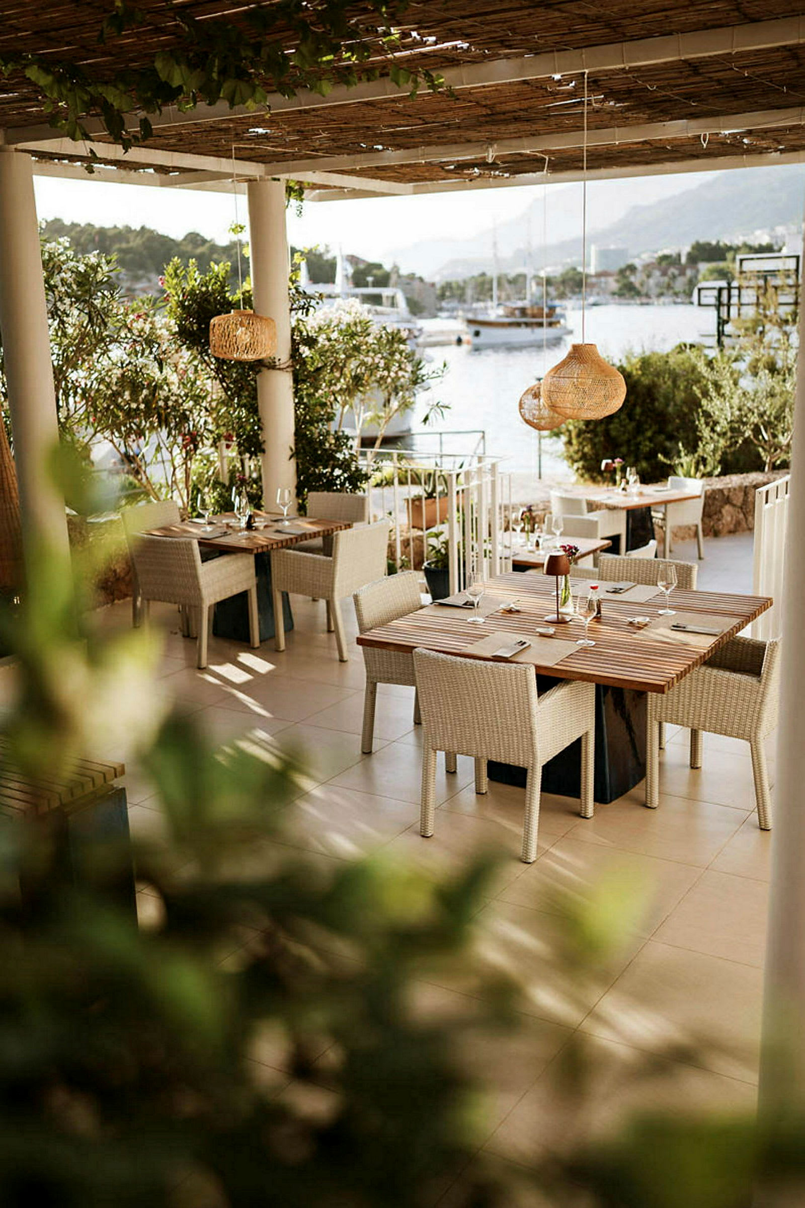En utomhus restaurang med bord och stolar under tak, nära vatten med båtar i bakgrunden. (AI-genererad text)