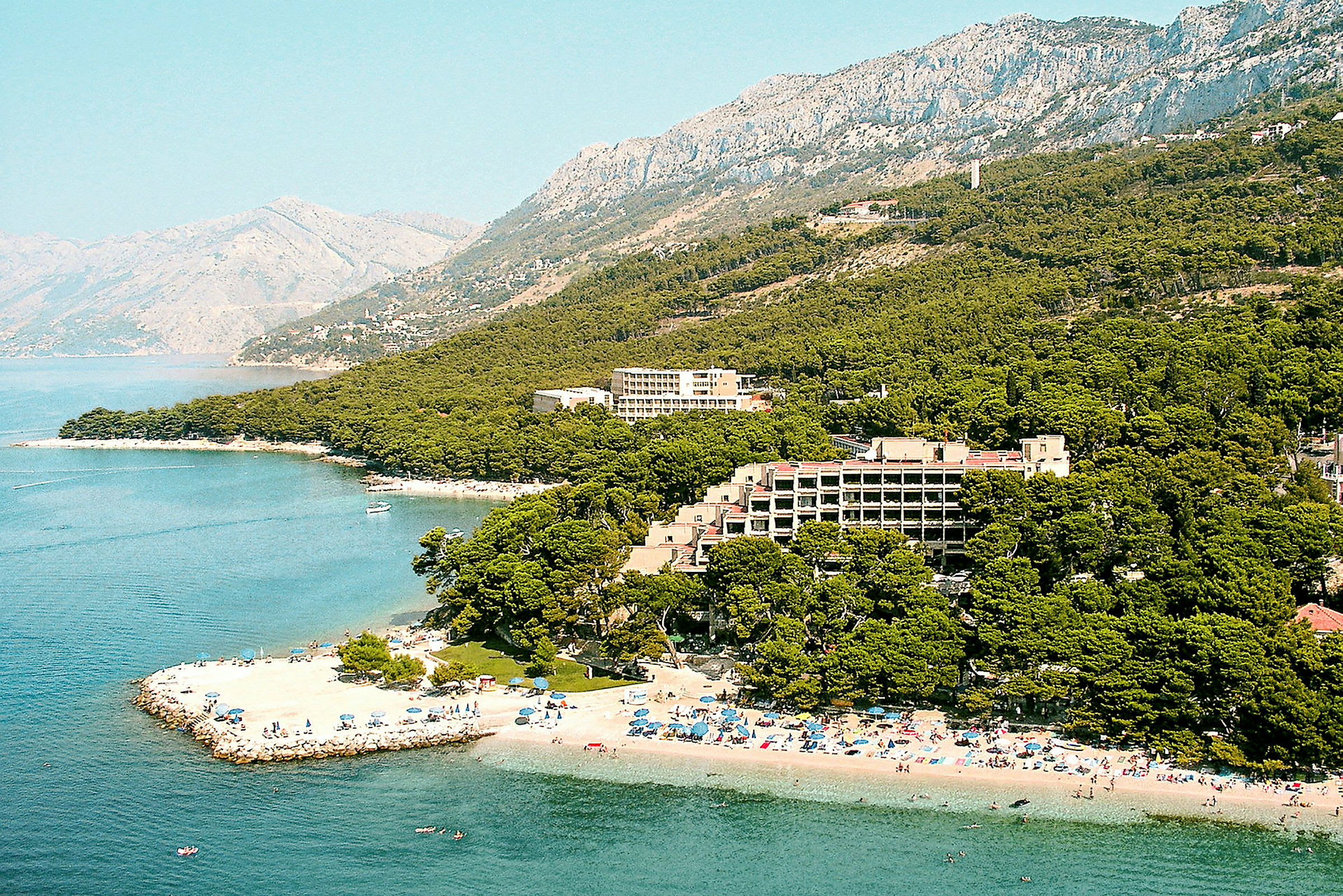 Strand, hotell och grönska vid en kust med berg i bakgrunden. (AI-genererad text)