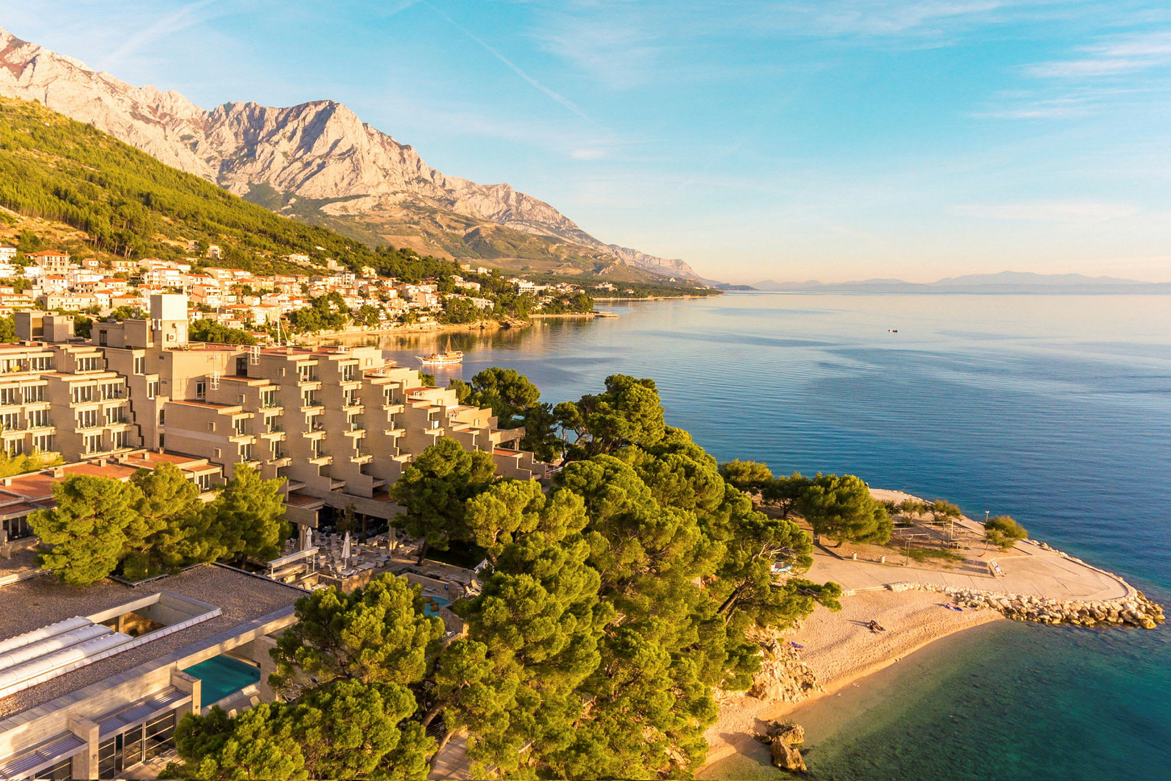 Utsikt över en kustlinje med hotellbyggnader, tallar, strand och hav i solnedgången. (AI-genererad text)
