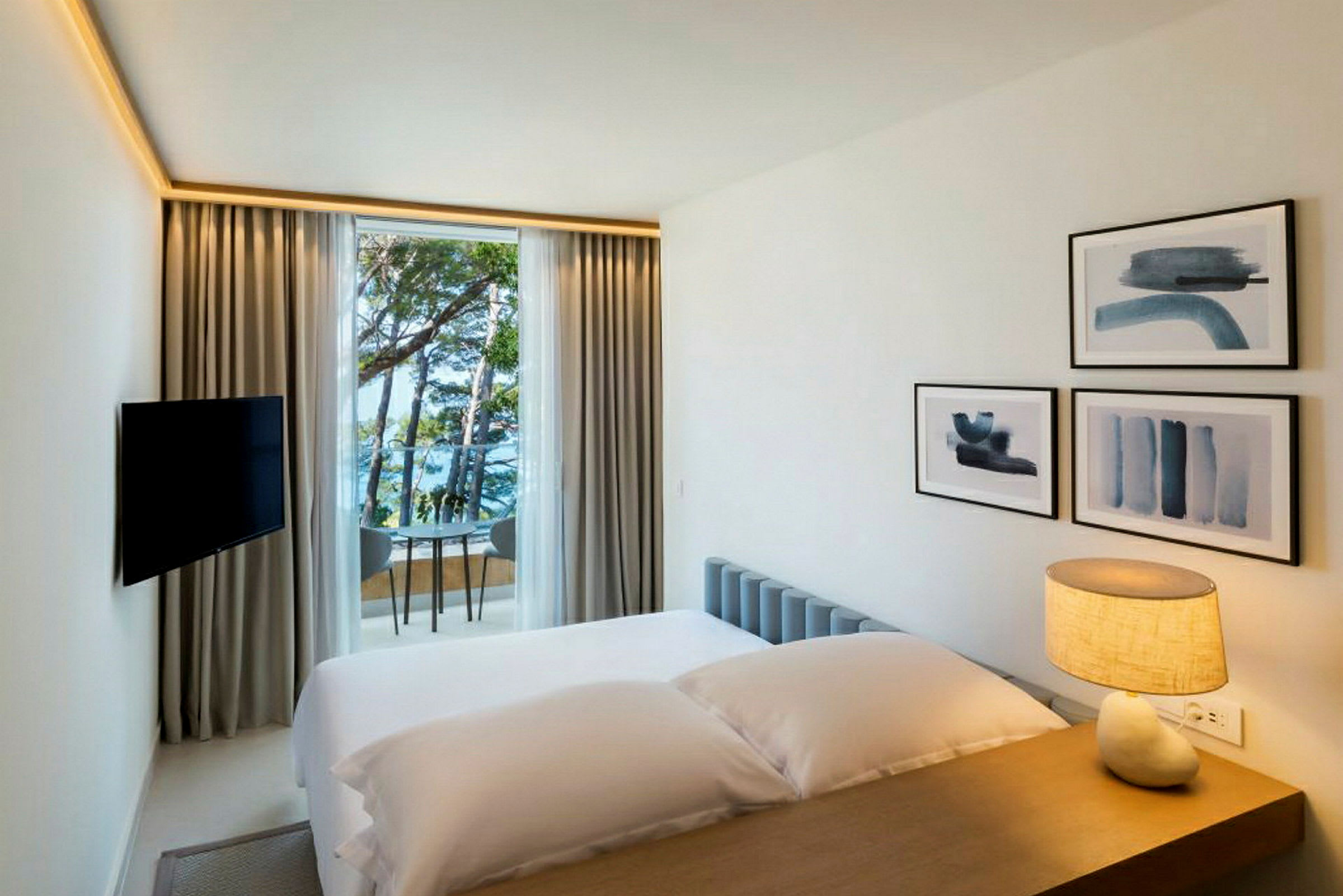 Ett hotellrum med en säng, en lampa på bordet, väggdekorationer och ett fönster som vetter mot havet och träd. (AI-genererad text)