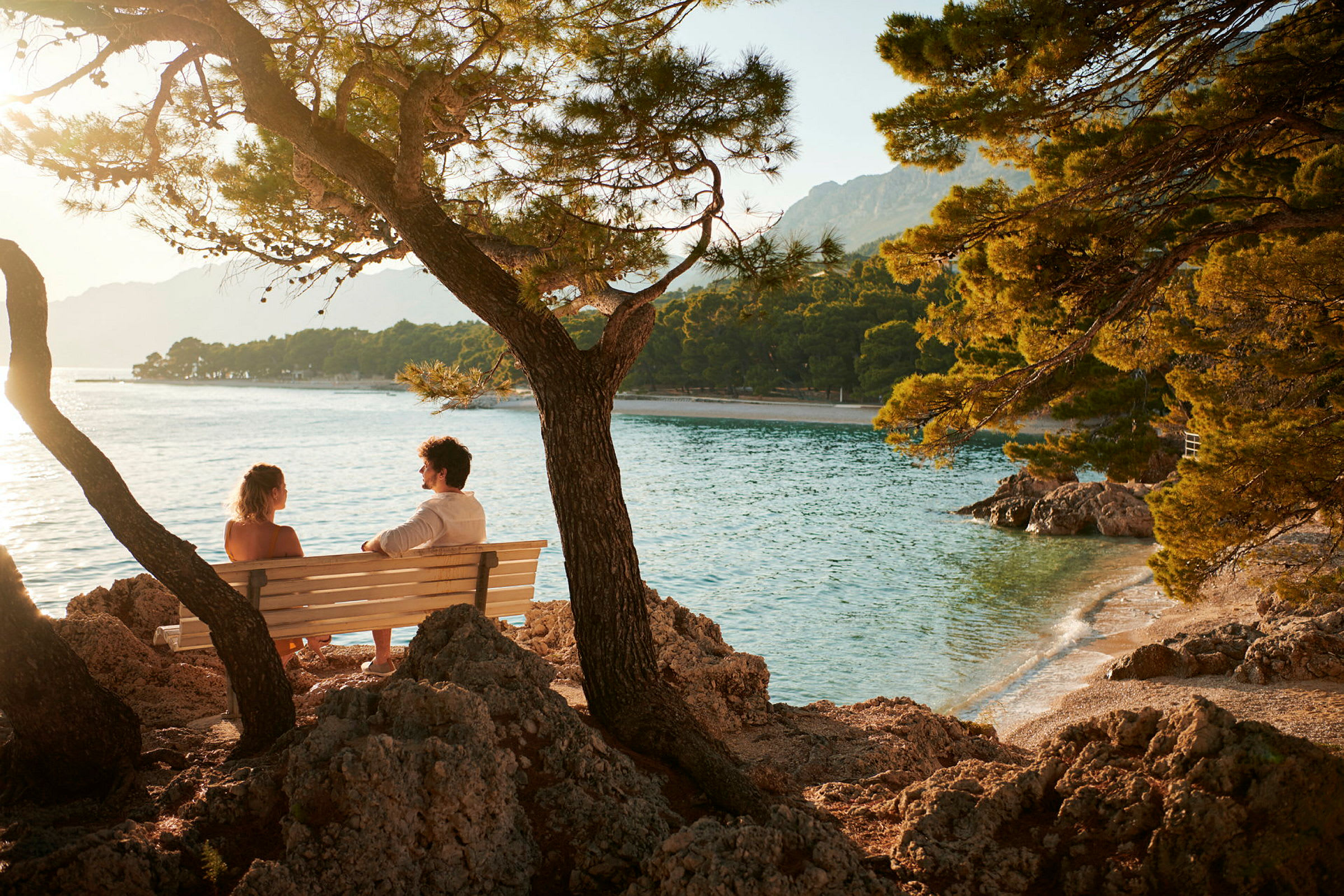 Två personer sitter på en bänk vid kustlinjen med utsikt över havet och träd. (AI-genererad text)