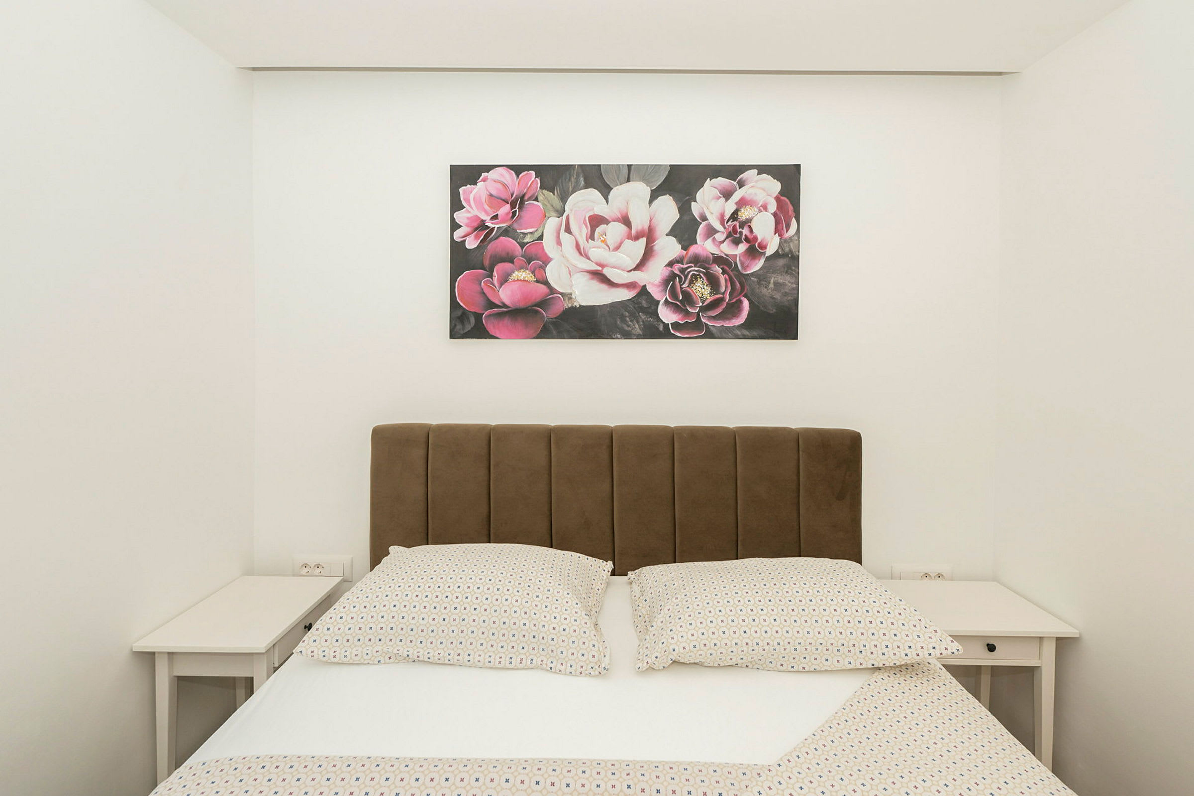 Säng med brun huvudgavel, två sängbord och en tavla med blommotiv ovanför. (AI-genererad text)