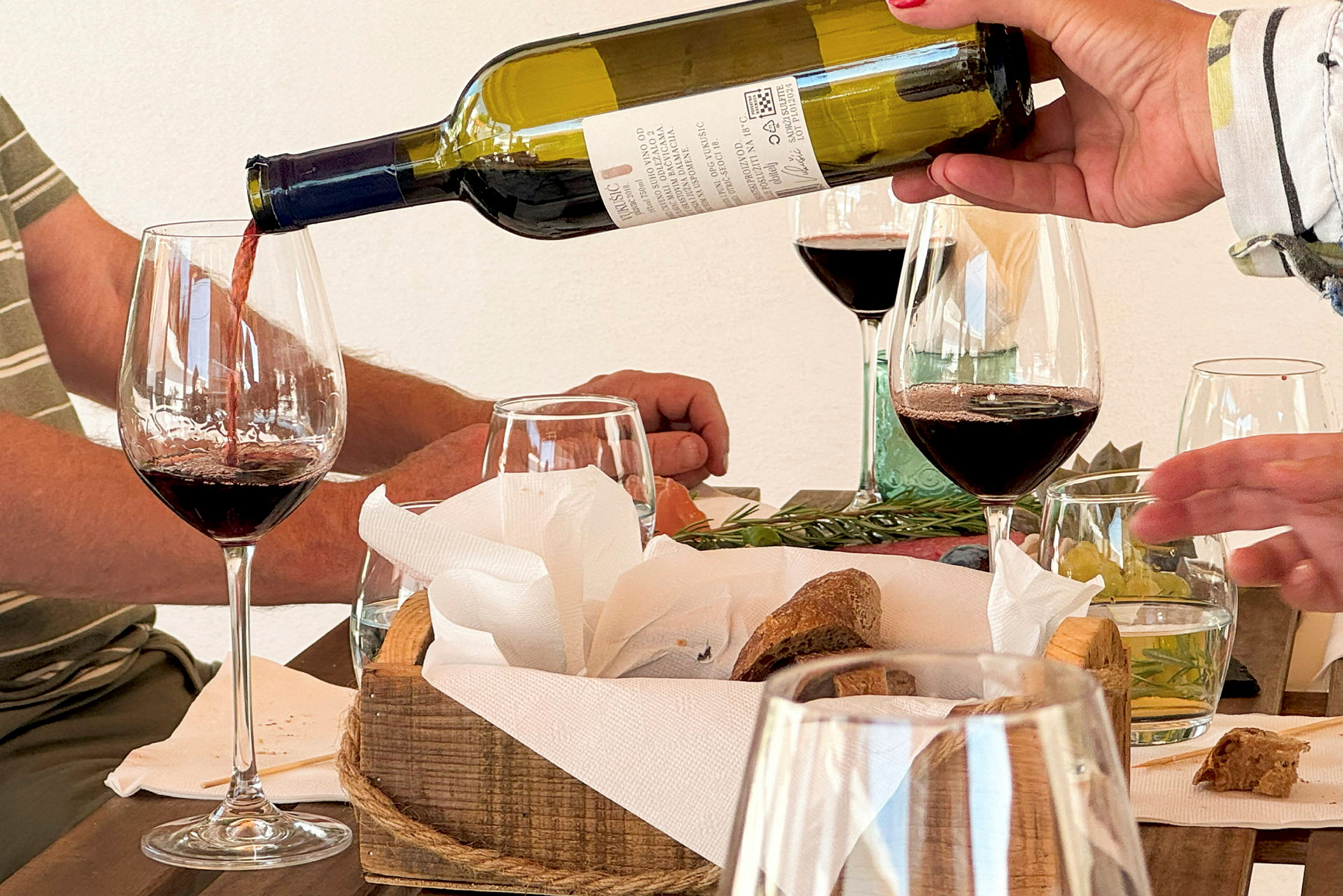 En person häller upp ett glas rött vin vid ett bord med bröd och tillbehör. (AI-genererad text)