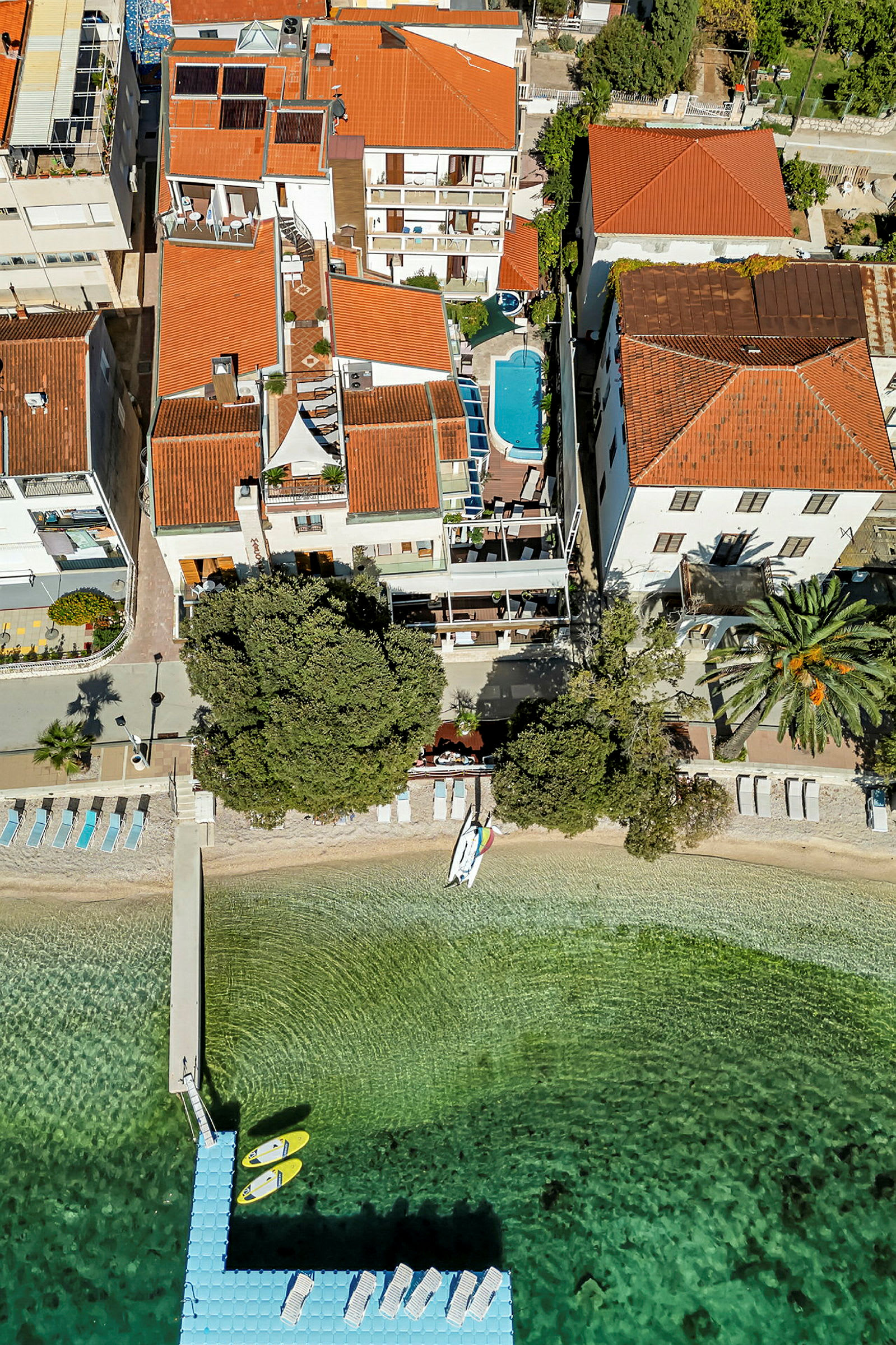 Flygbild över en byggnad med takterrass, en pool och en brygga med kajaker vid en strand. (AI-genererad text)