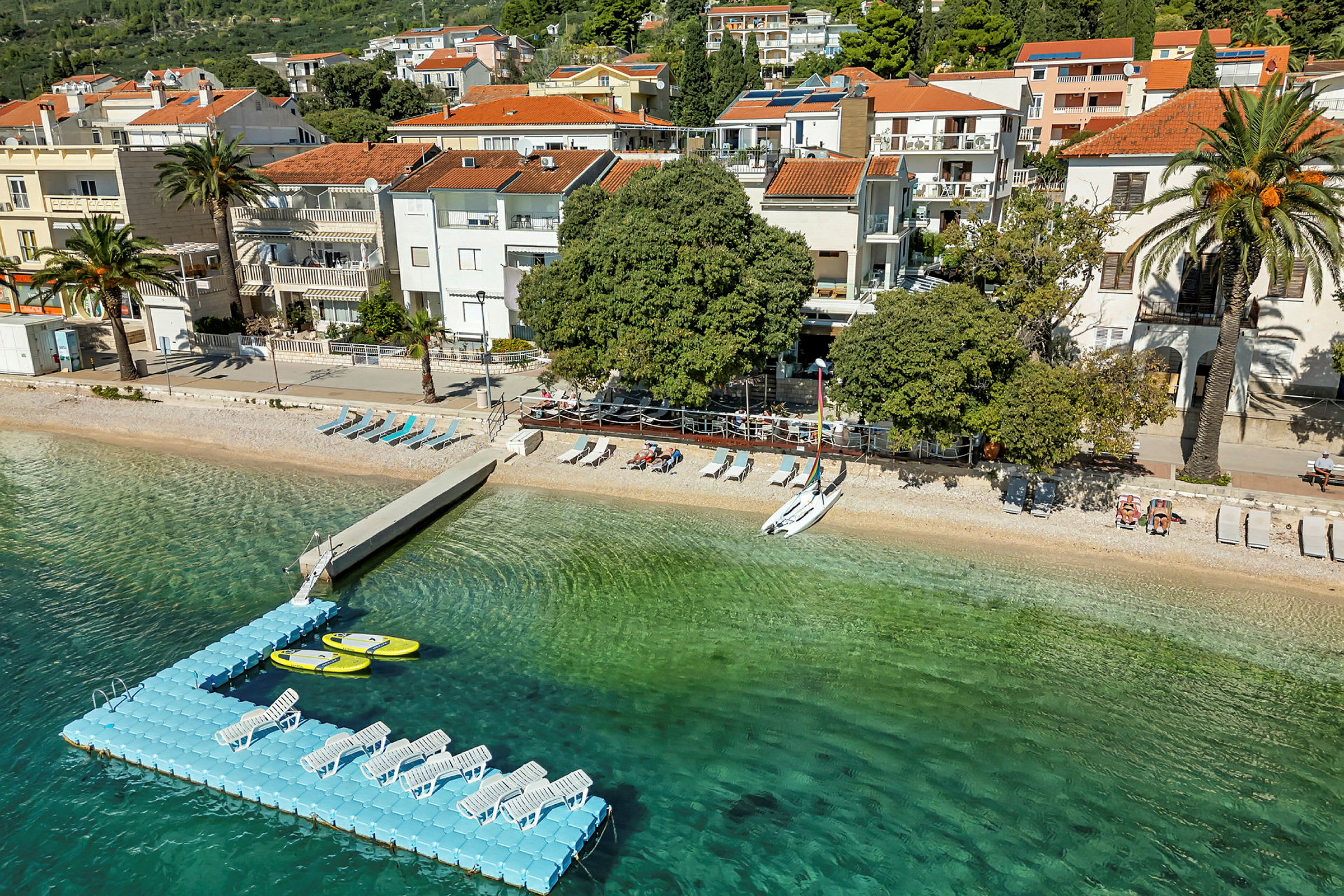 Strand med solstolar och flytbrygga, med byggnader i bakgrunden. (AI-genererad text)