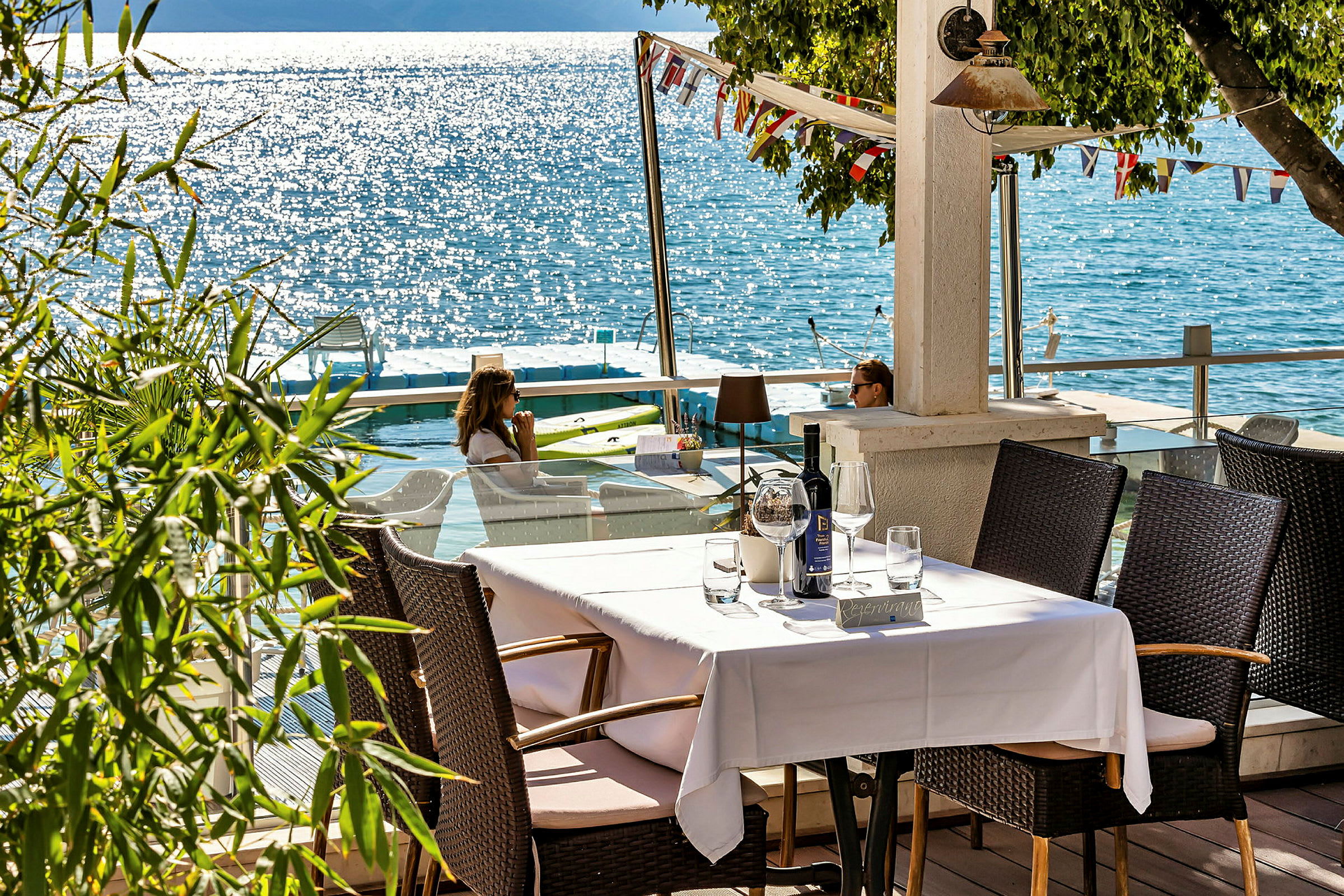 Ett dukat bord på en terrass med utsikt över en solglittrande sjö. (AI-genererad text)