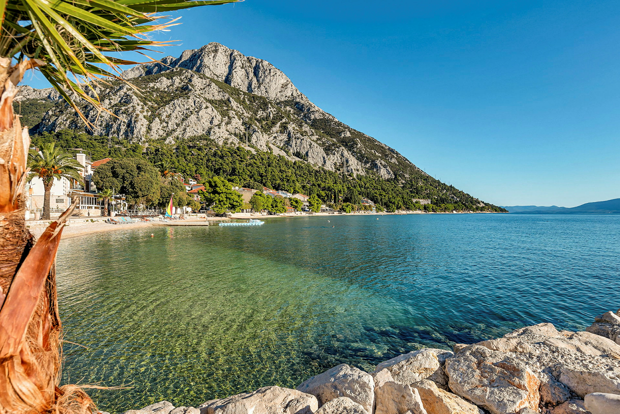 Bilden visar en strand med klart vatten, omgiven av grönska och berg i bakgrunden. (AI-genererad text)