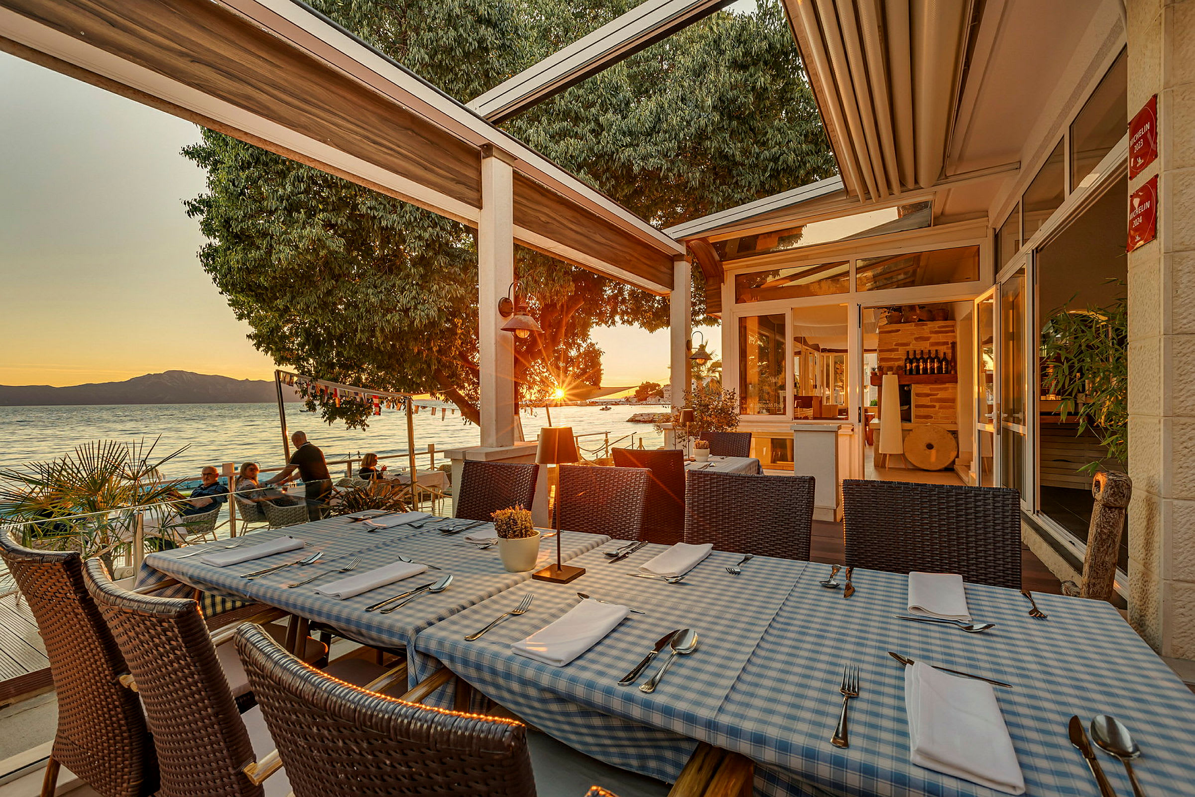 Restaurangbord med blåvitrutiga dukar på en terrass vid havet under solnedgången. (AI-genererad text)