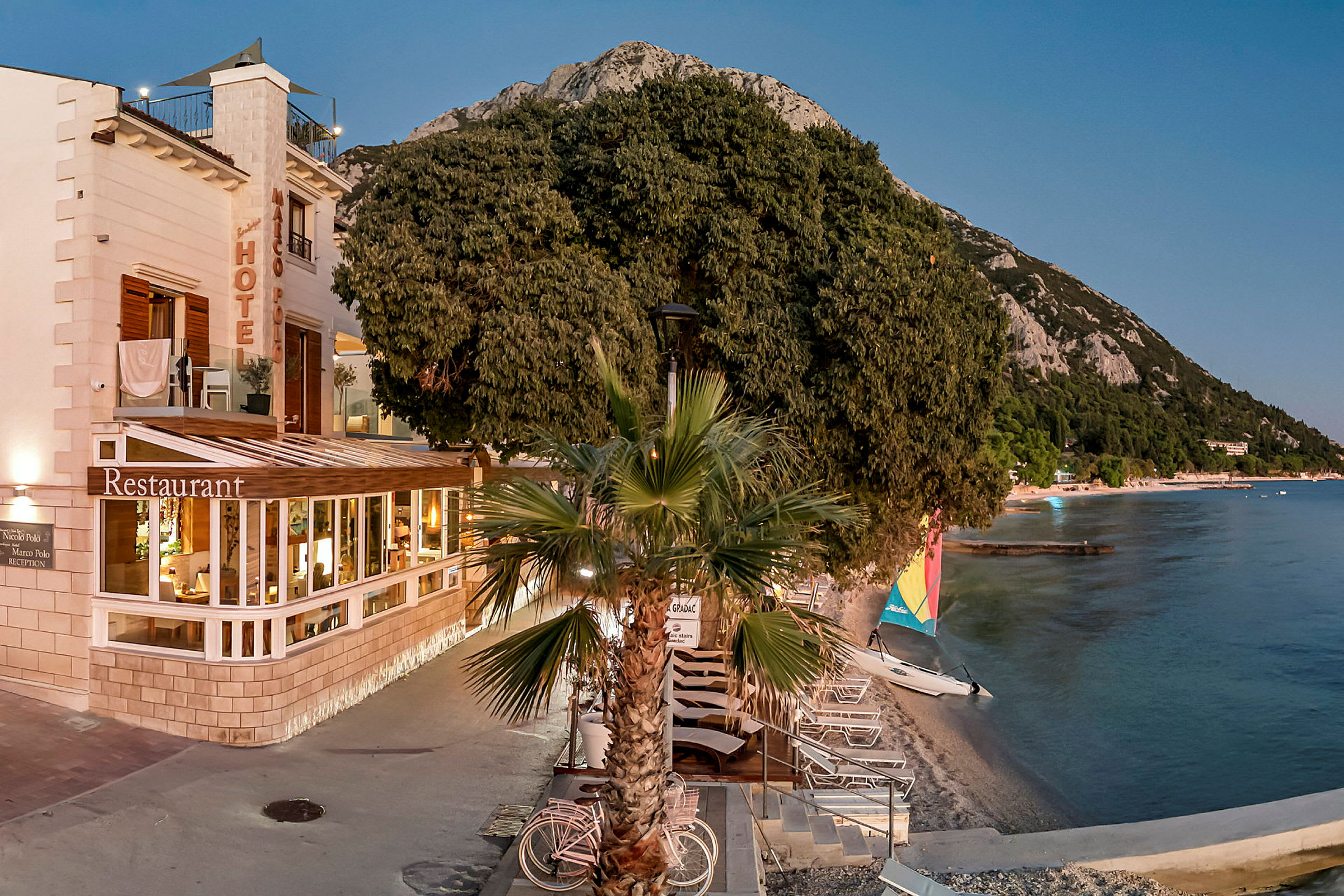 Bild av ett hotell vid stranden, med en restaurang och solstolar längs vattenkanten. (AI-genererad text)
