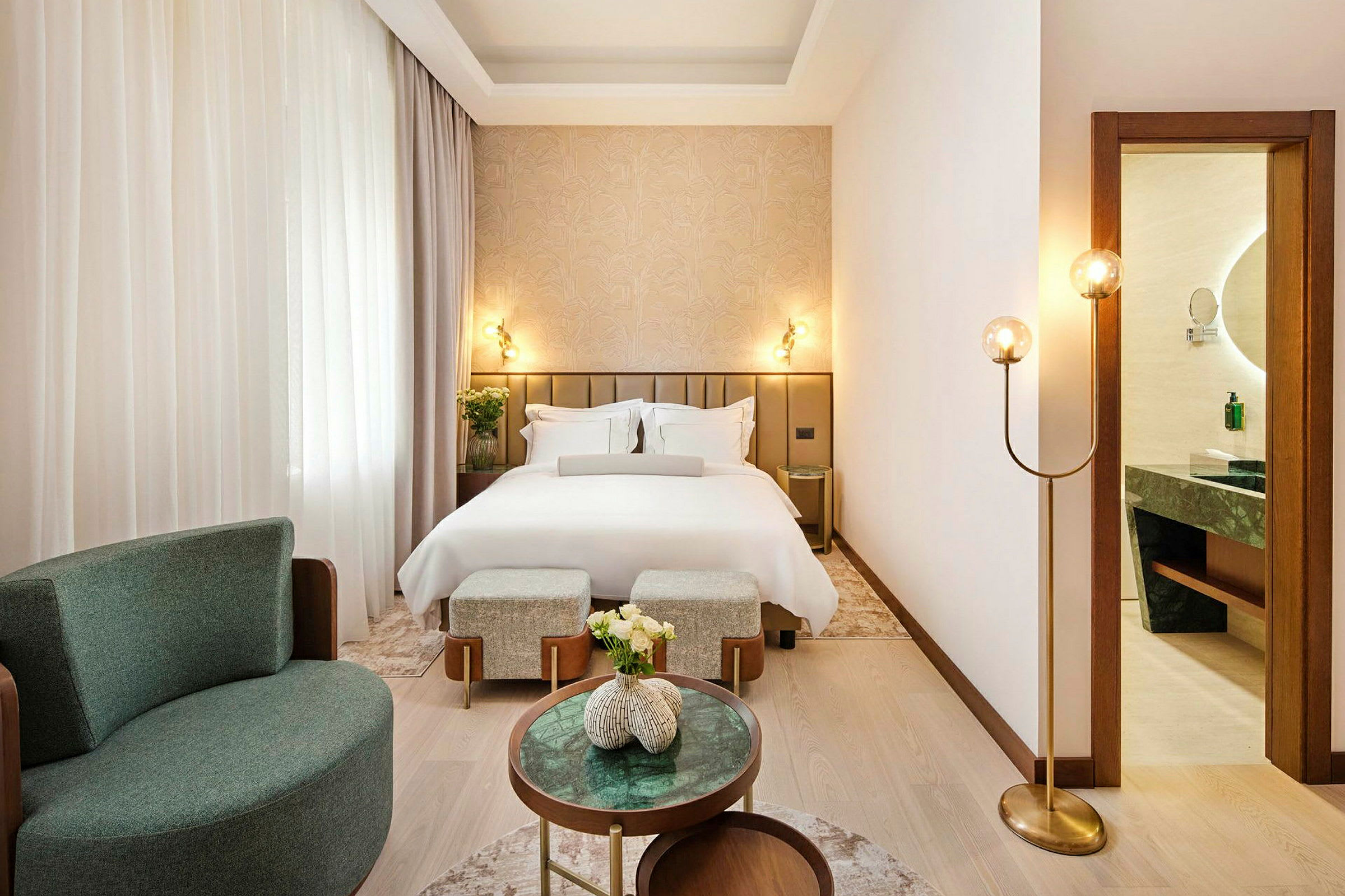 Ett elegant hotellrum med en bäddad säng, fåtölj och små bord. Ett badrum syns genom en öppnad dörr. (AI-genererad text)