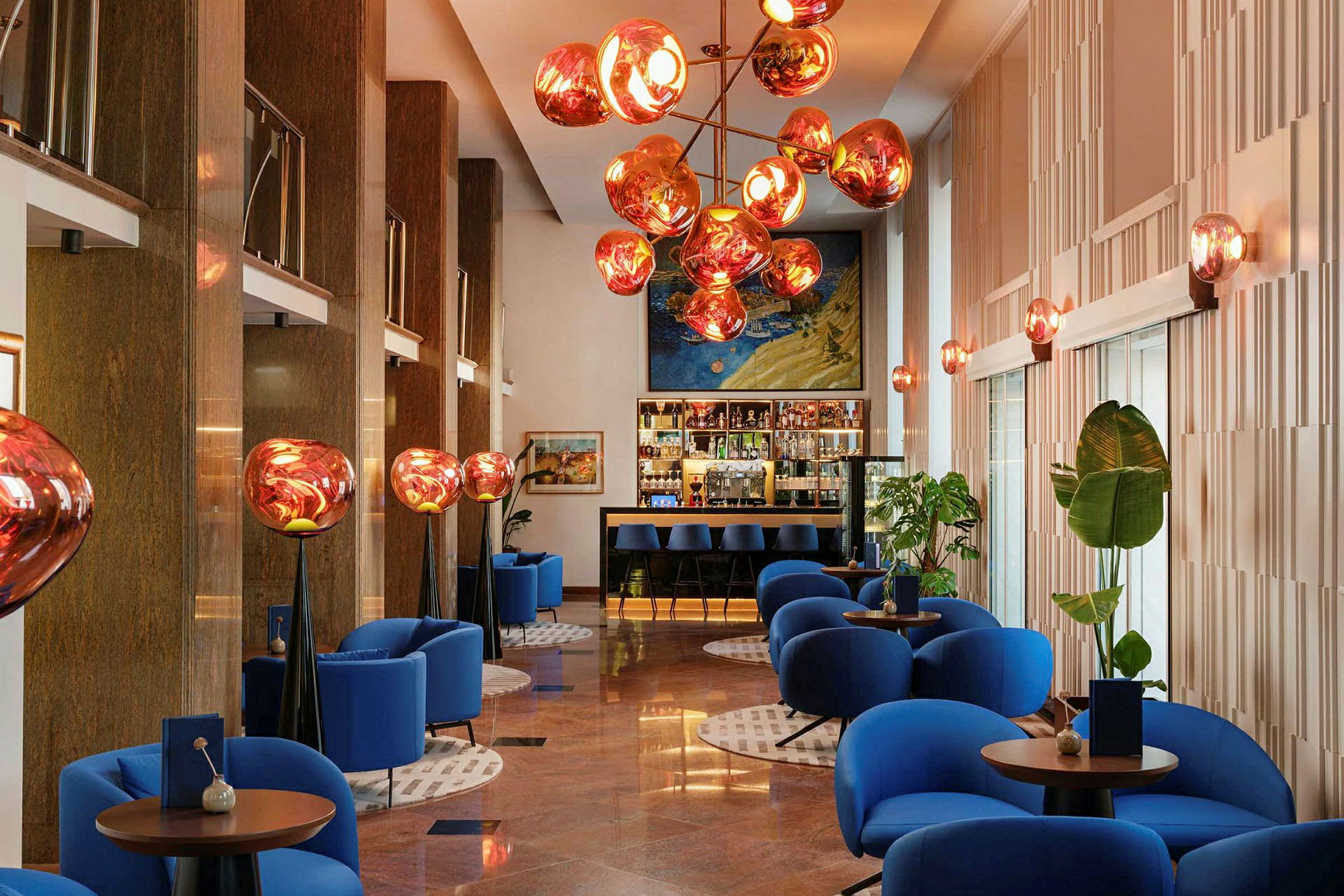 Ett modernt inrett hotelllobby med blå fåtöljer, runda bord och en dekorativ belysning i taket. (AI-genererad text)