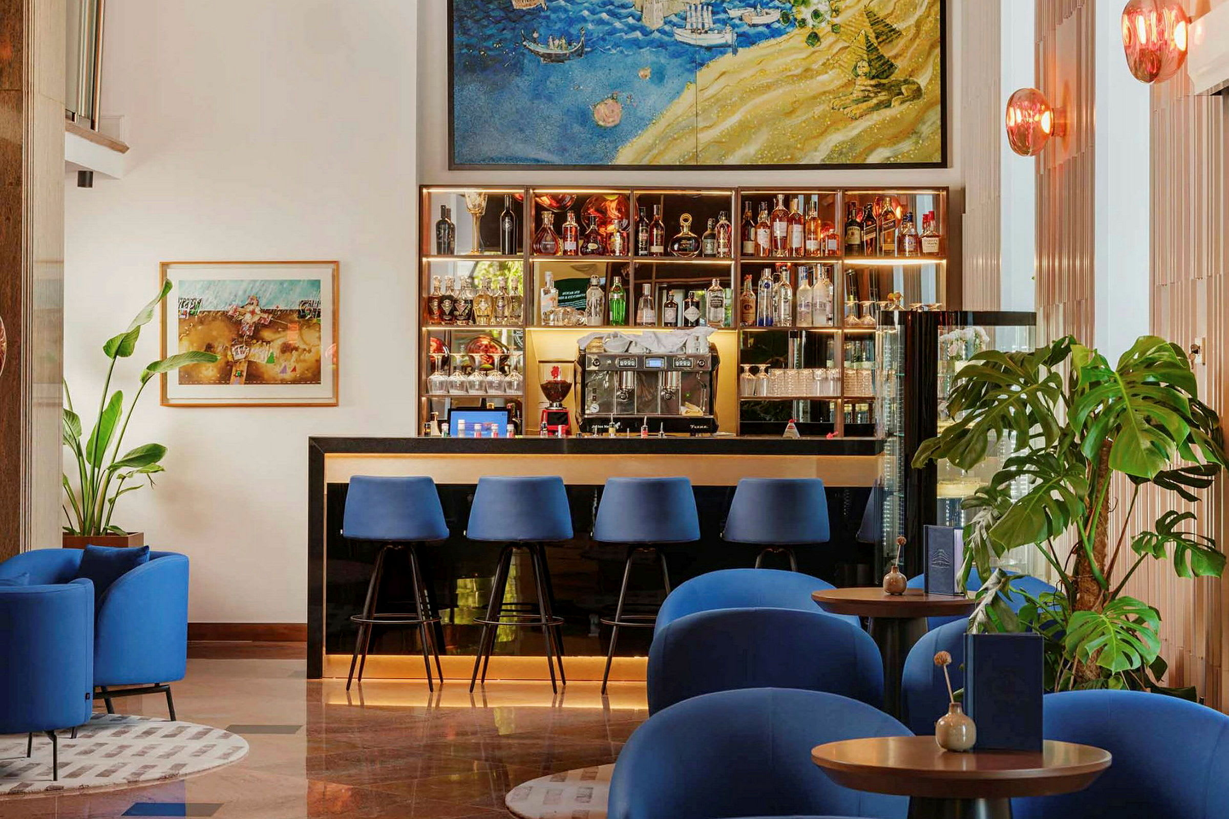 En hotellbar med blåa stolar vid disken samt blå fåtöljer framför små bord. Väggdekorationer och växter finns i bakgrunden. (AI-genererad text)