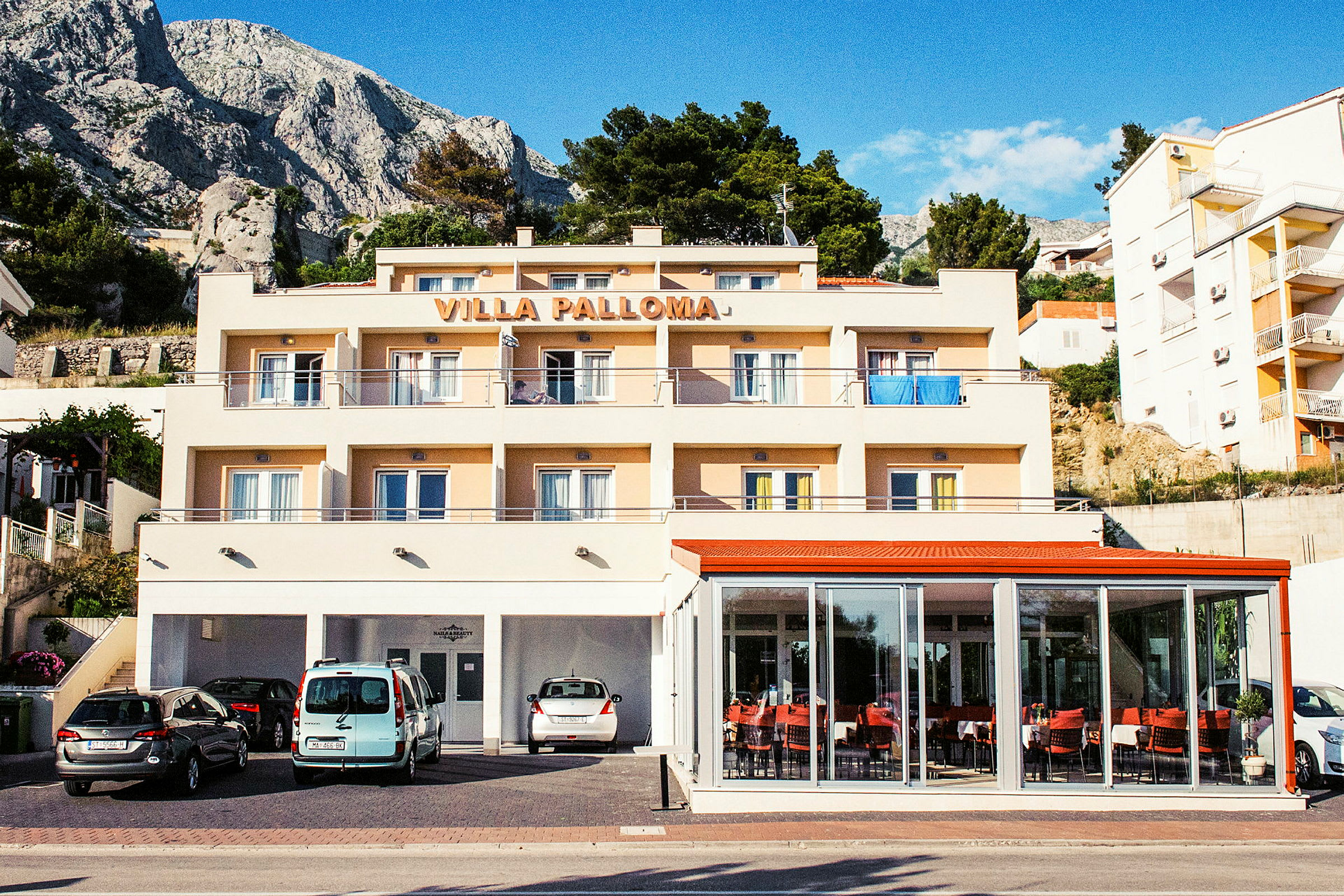 En byggnad med texten 'Villa Palloma' på toppen, med balkonger och parkerade bilar framför. (AI-genererad text)