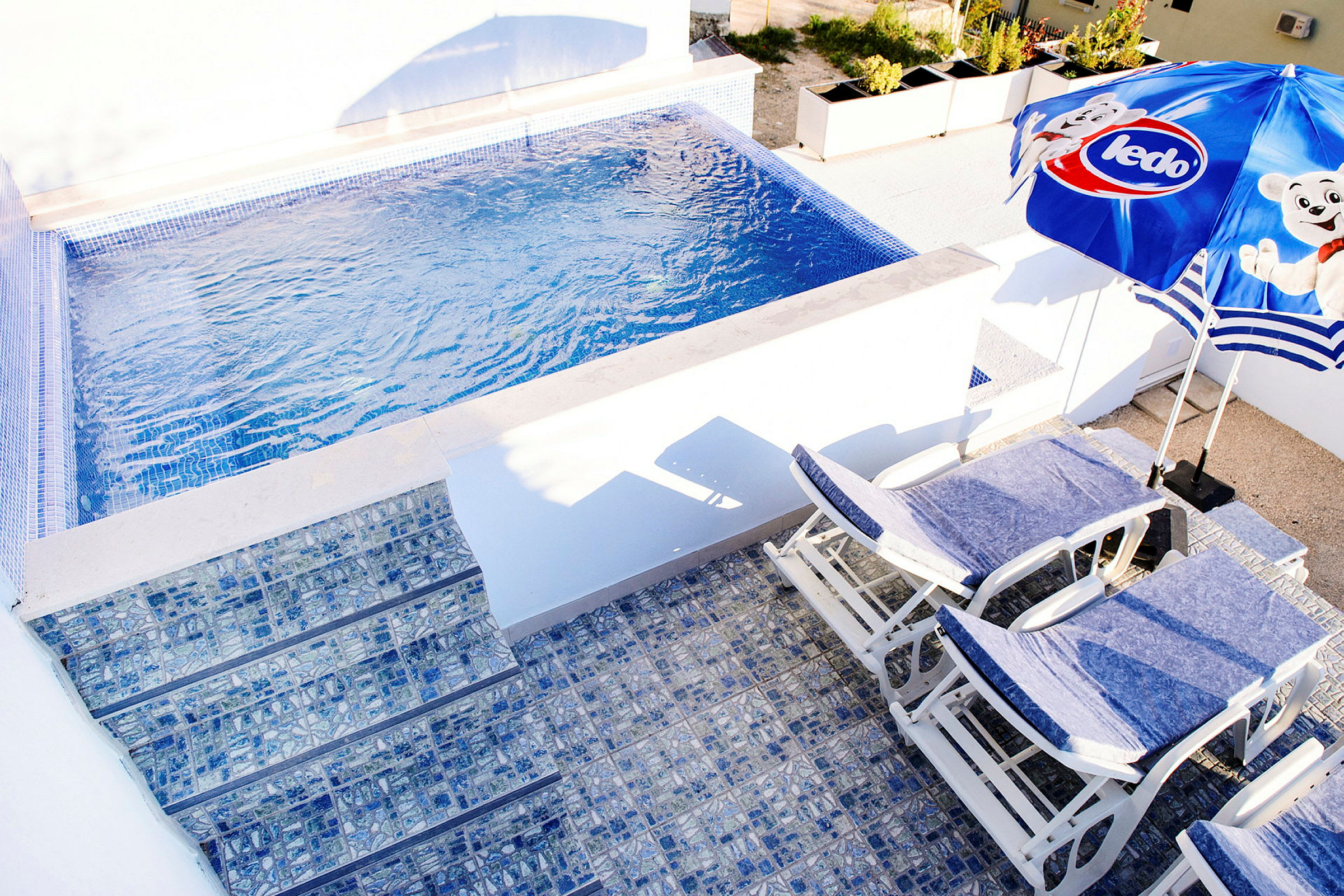 Bild av en liten pool med blå mosaik och solstolar samt en parasoll. (AI-genererad text)