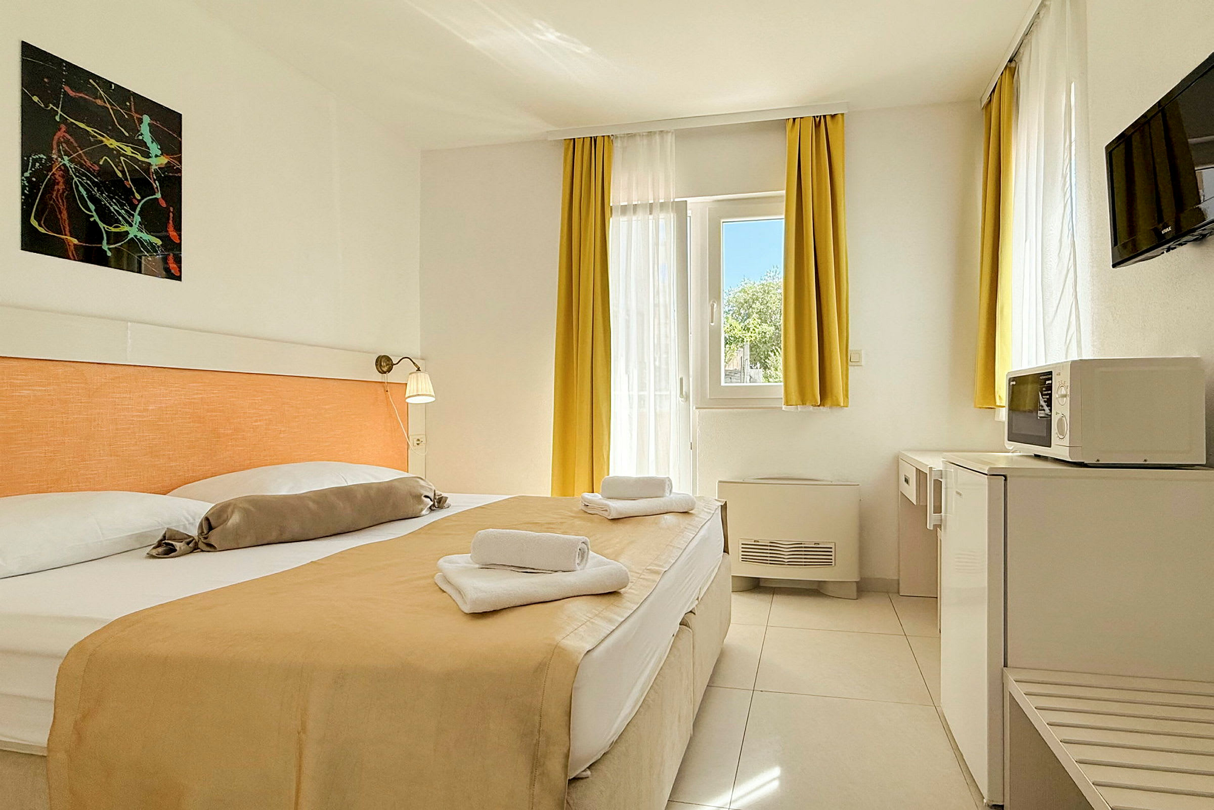 Ett hotellrum med en dubbelsäng, ljusa gardiner och möbler samt ett fönster som släpper in dagsljus. (AI-genererad text)