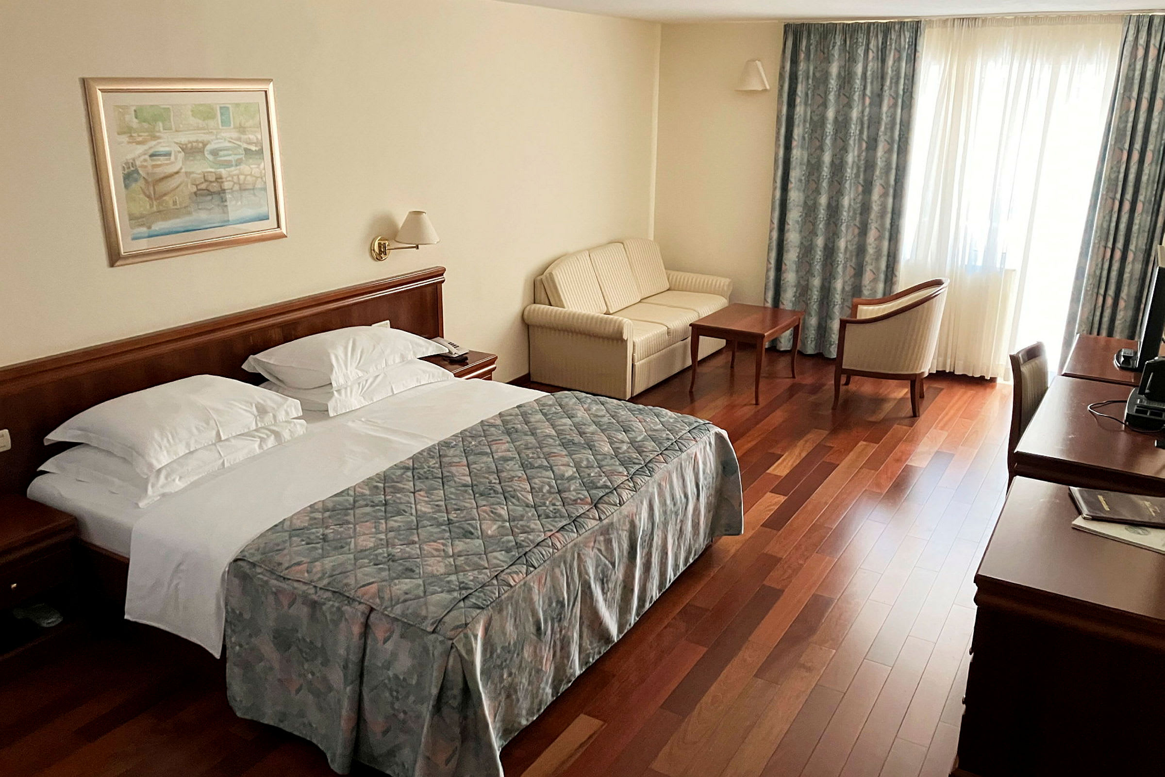 Ett hotellrum med en dubbelsäng, soffa, träbord och stolar samt ett skrivbord med TV. (AI-genererad text)