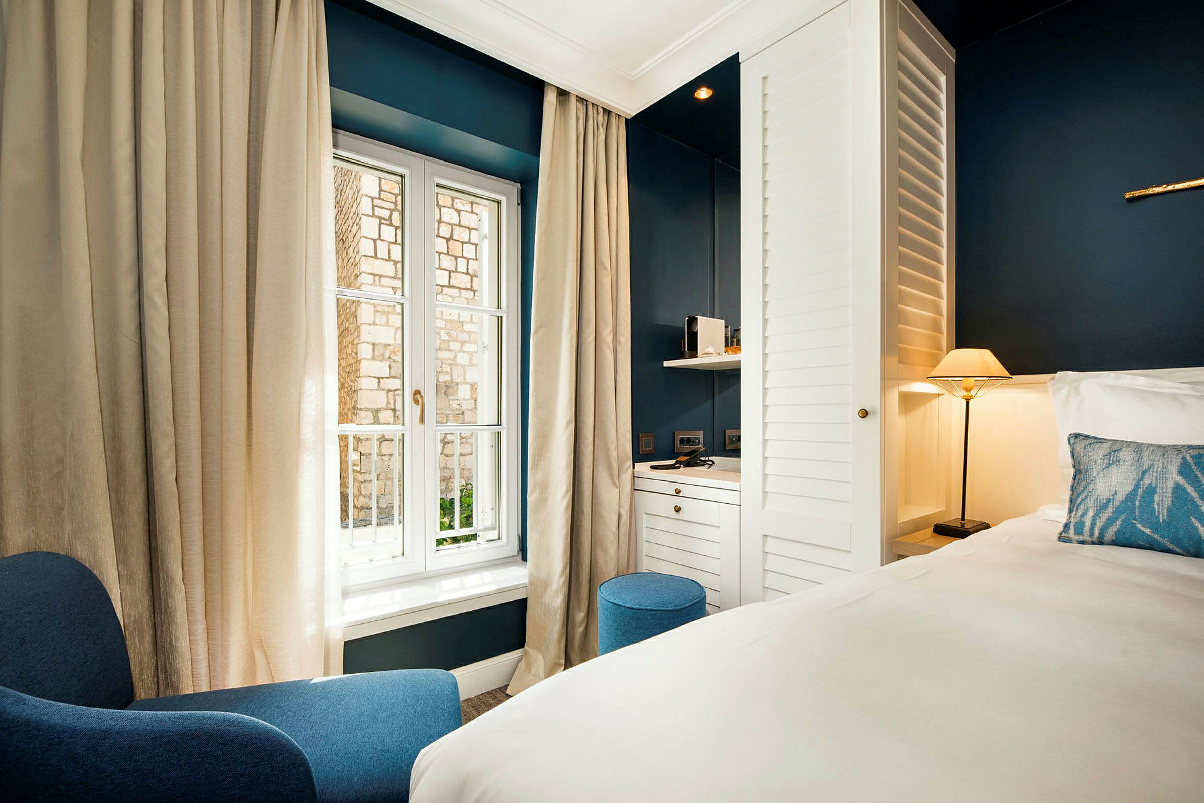 Ett hotellrum med moderna möbler inkluderande en blå fåtölj, en säng med vitt sängkläder, en liten lampa och ett skrivbord vid ett fönster. (AI-genererad text)