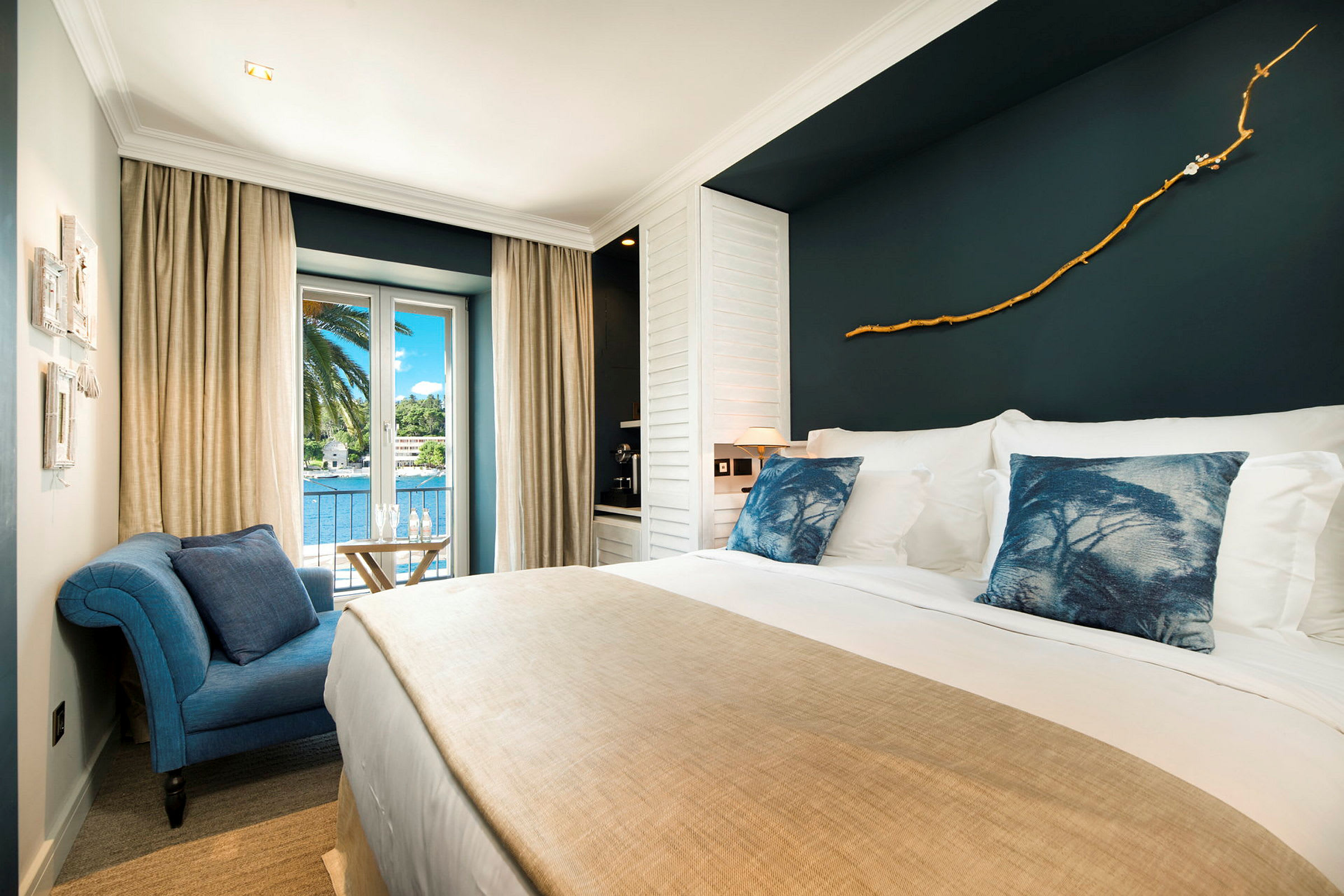 Ett elegant hotellrum med stor säng, blå prydnadskuddar, beige sängöverkast och dekorerad mörk vägg. Rummet har ett fönster med utsikt över palmträd och vatten. (AI-genererad text)