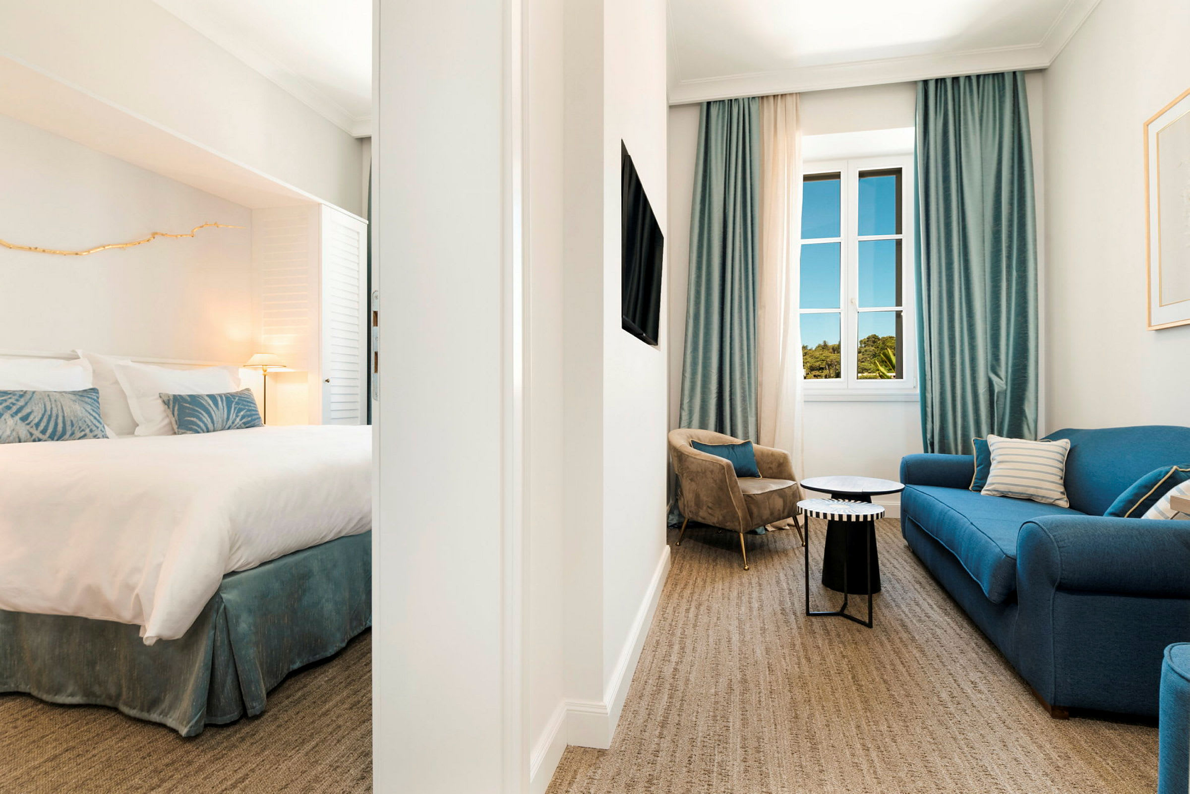 En hotellsvit med sovrum och vardagsrumsdel, inredd med en blå soffa, fåtöljer och fönster med blå gardiner. (AI-genererad text)