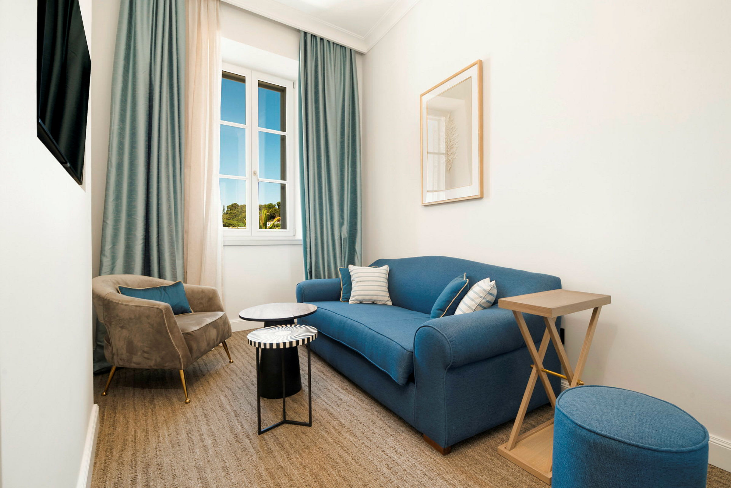 Ett ljust vardagsrum med en blå soffa, ett fåtölj i beige, små bord och en fönsterutsikt mot grönska utomhus. (AI-genererad text)