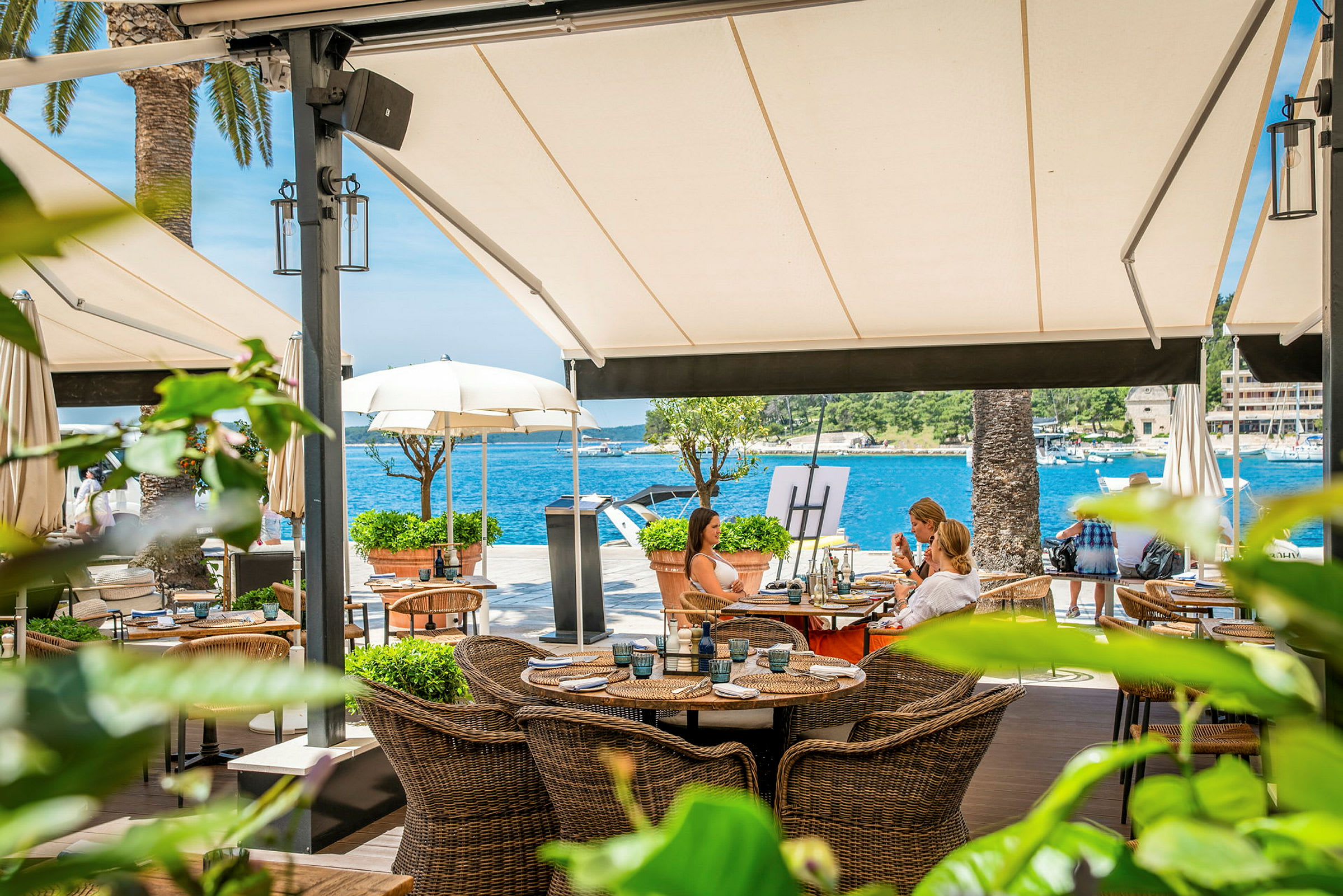 En restaurangterrass vid vattnet med korgmöbler och utsikt över havet under stora markiser. (AI-genererad text)