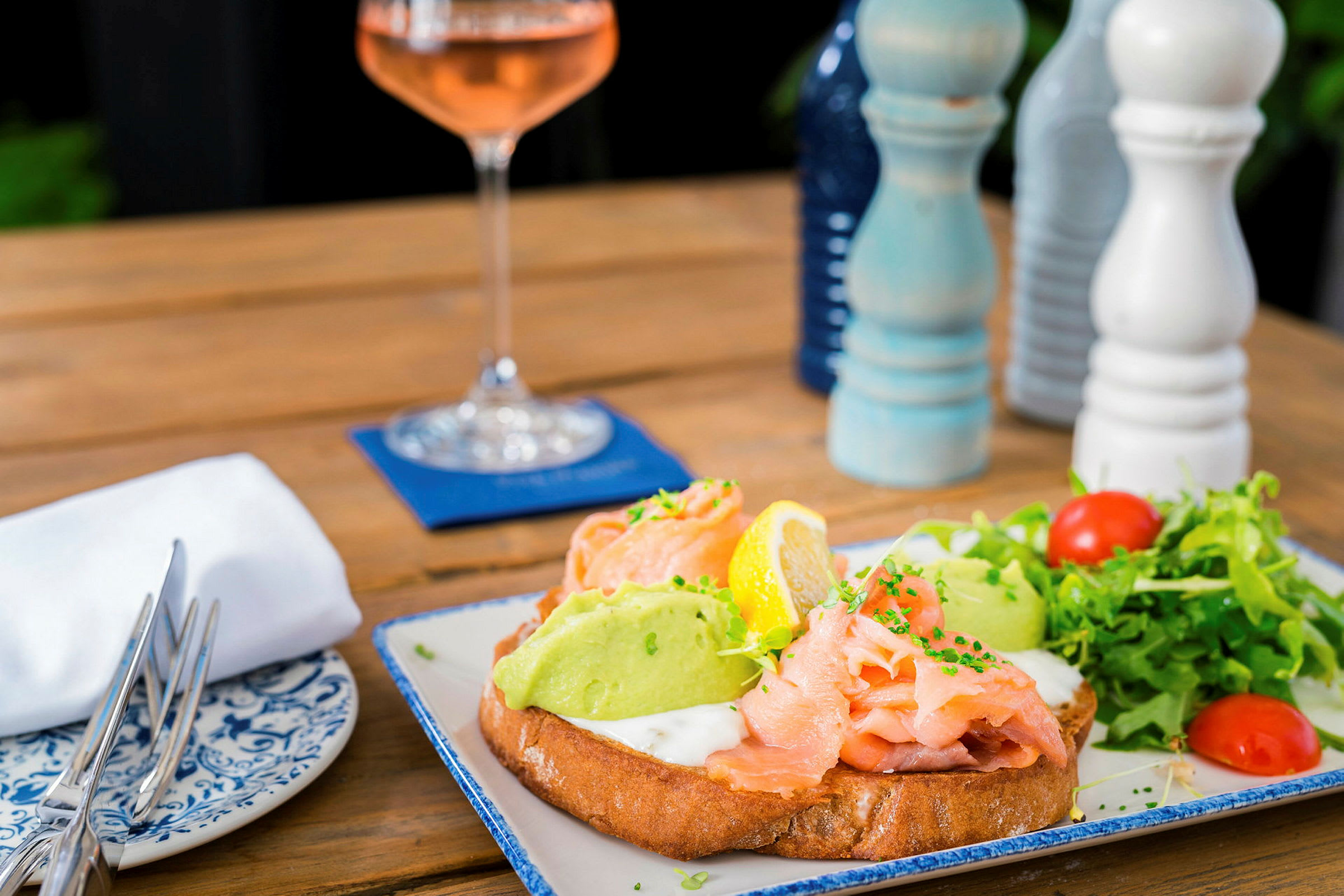 En tallrik med mat: smörgås med lax, avocado och grönsaker. I bakgrunden finns en vinglas och kryddkvarnar. (AI-genererad text)