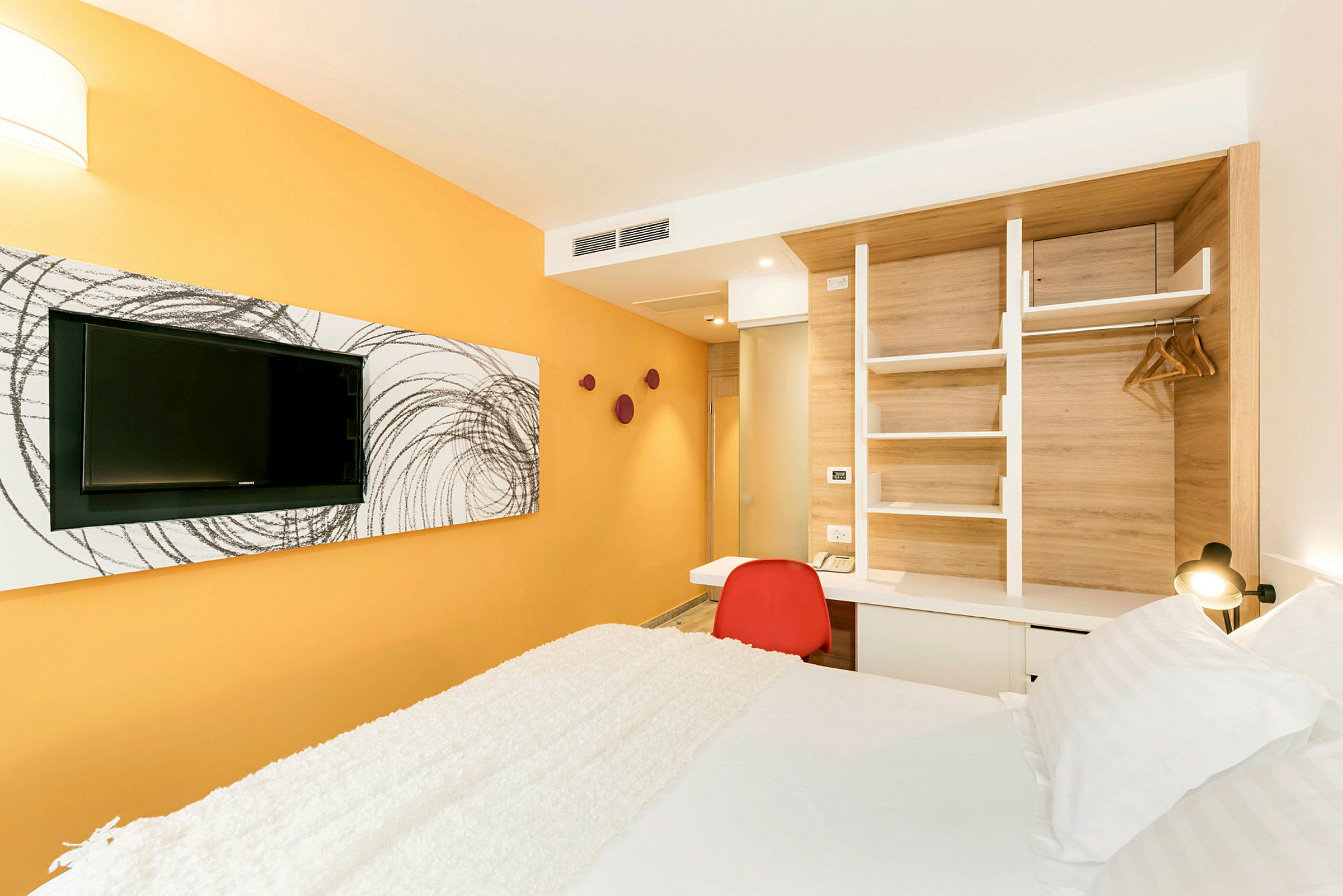 Ett hotellrum med gul vägg, en TV på väggen, en röd stol vid ett skrivbord och en säng med vita sängkläder. (AI-genererad text)