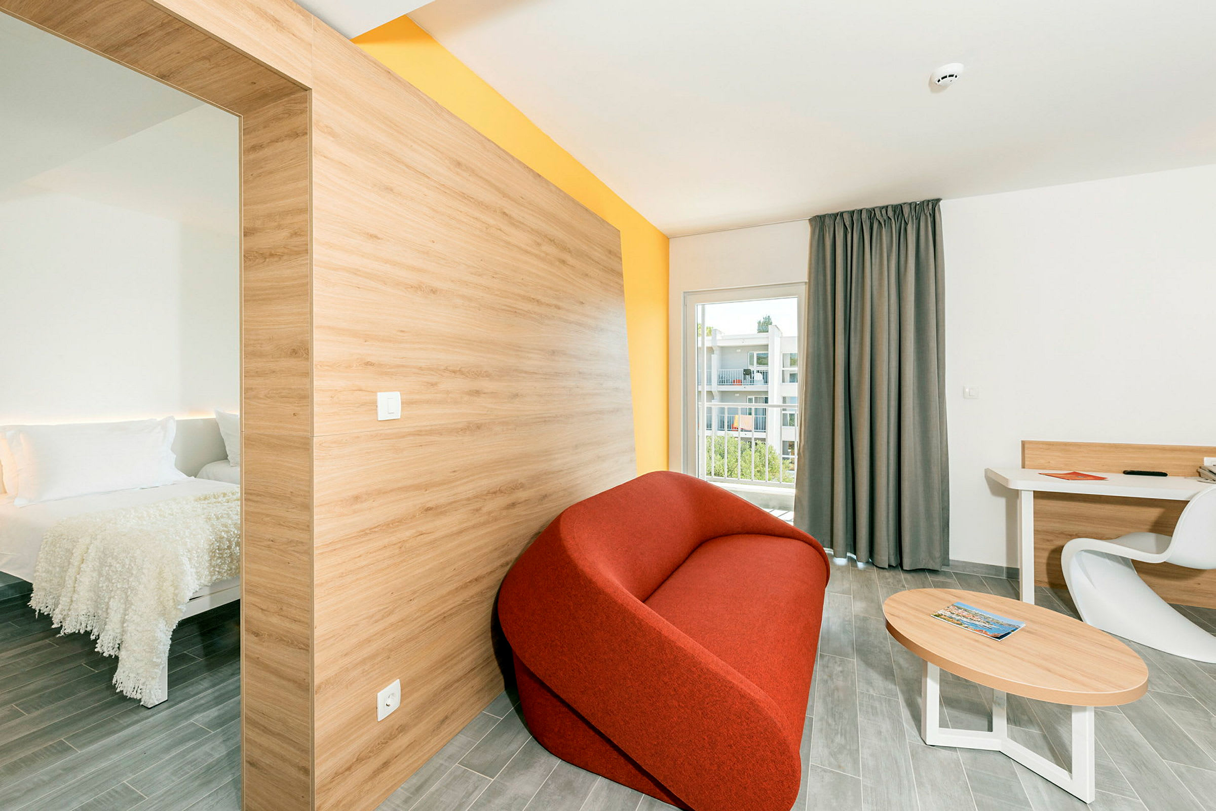 Ett modernt hotellrum med ett separat sovutrymme till vänster, en röd soffa, ett bord och en skrivplats med en stol, samt en balkong med utsikt. (AI-genererad text)