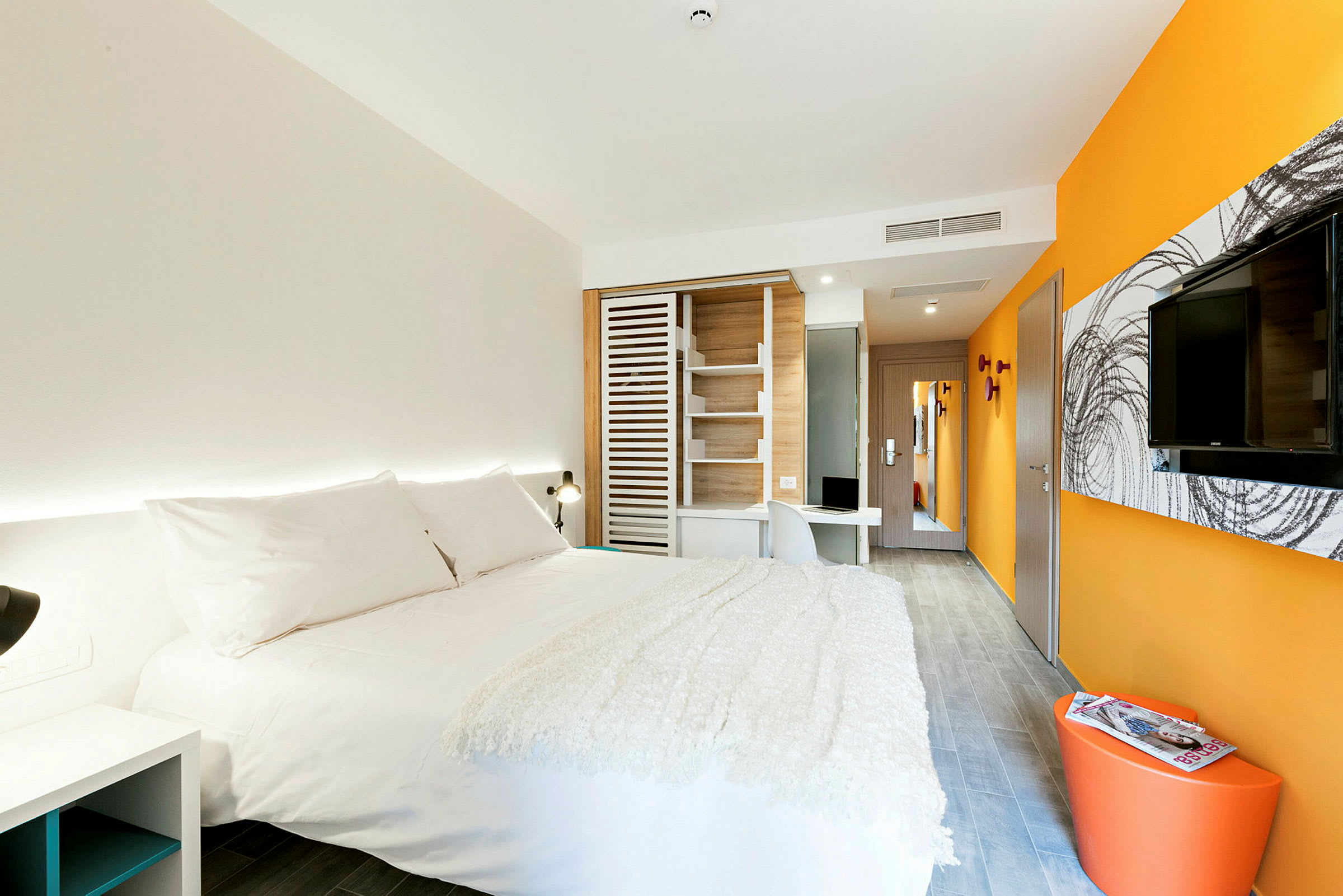 Ett modernt hotellrum med en stor säng klädd i vita sängkläder, orange och vita väggar, en väggmonterad TV och ett skrivbord. (AI-genererad text)