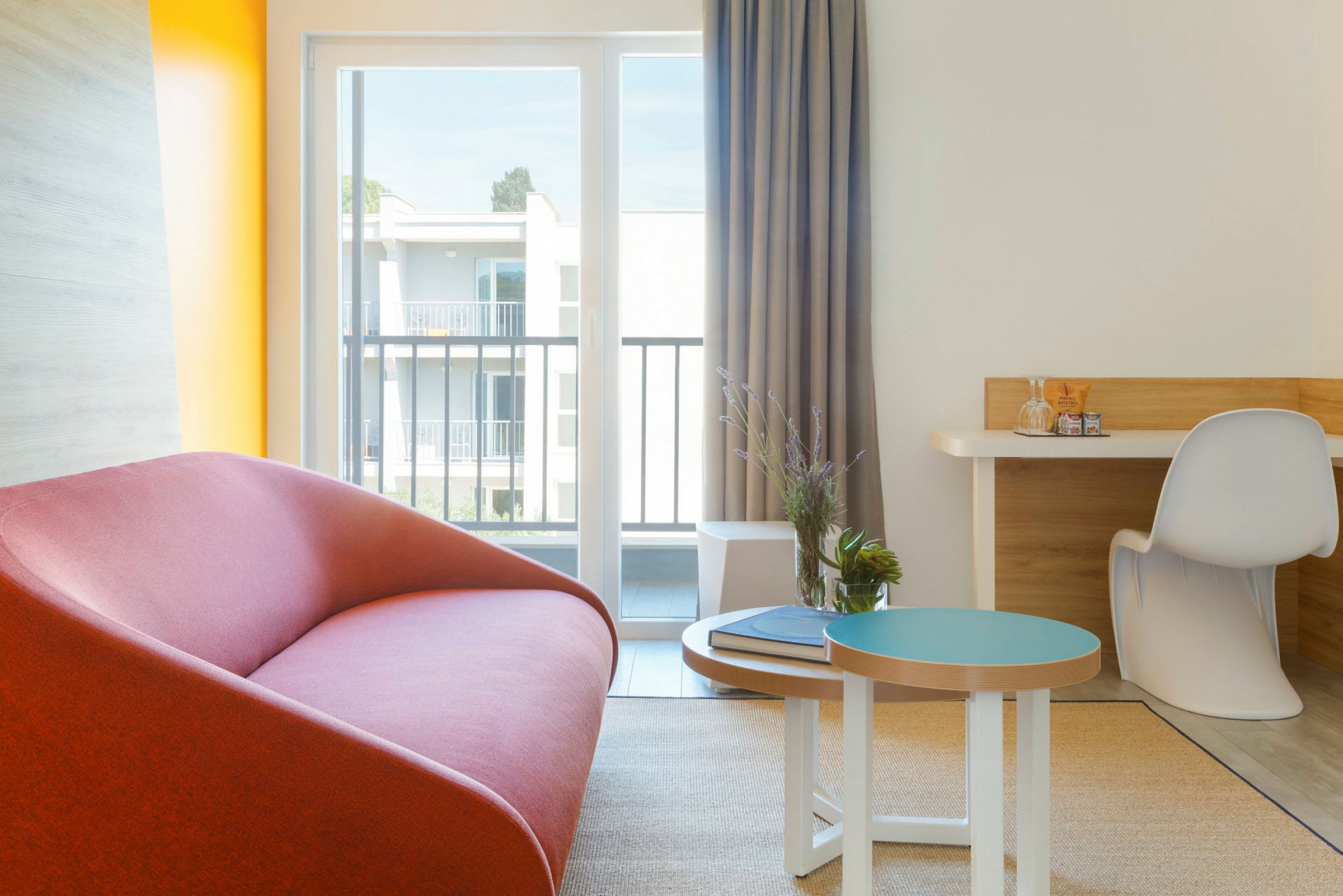 En modern inredd hotellrumsvrå med en röd soffa, ett litet bord med dekorationer och en skrivbordshörna vid ett stort fönster. (AI-genererad text)