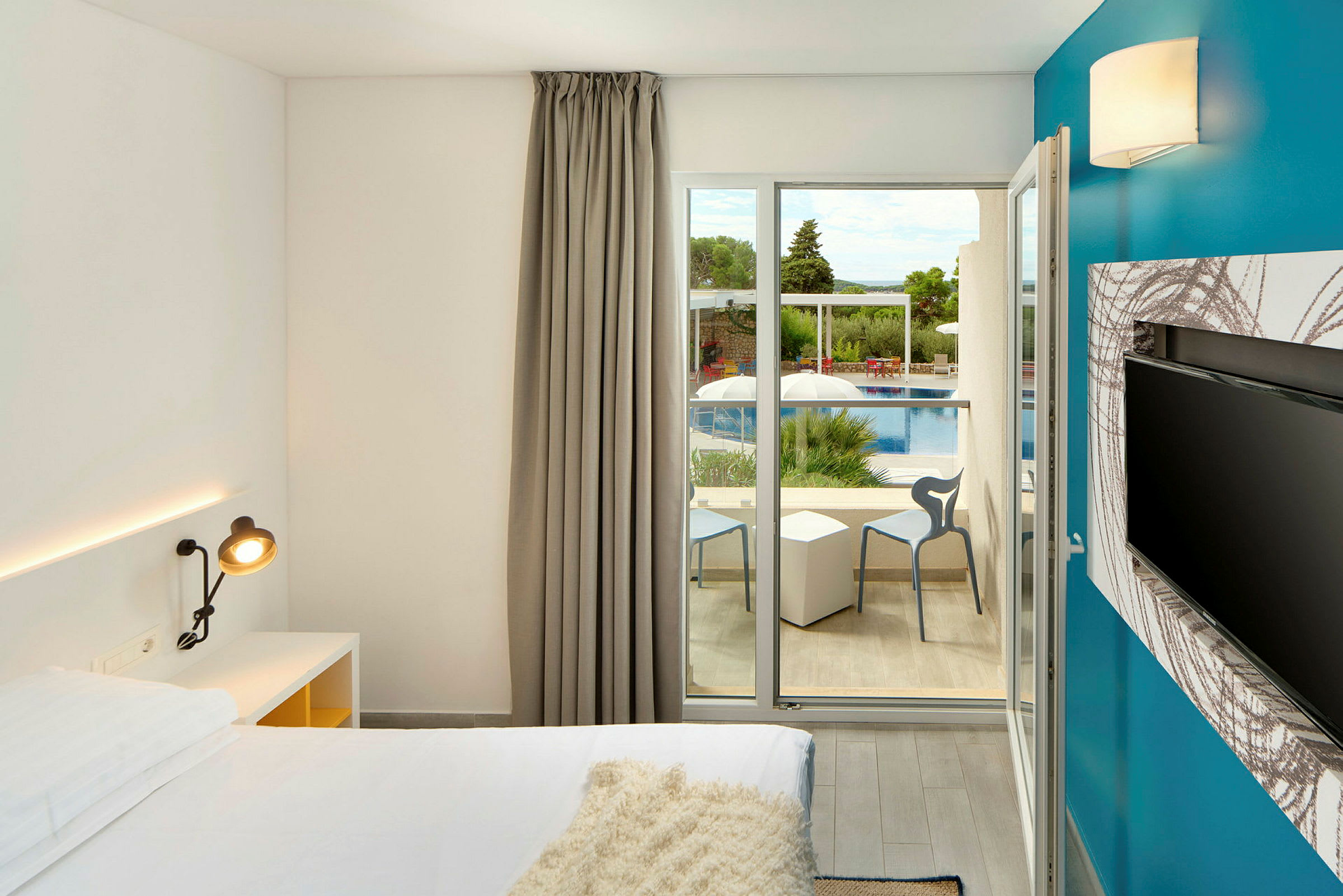 Ett modernt hotellrum med en säng, ett skrivbordslampa och en platt-TV. Fönsterdörrar leder ut till en balkong med utsikt över en pool och gröna träd. (AI-genererad text)