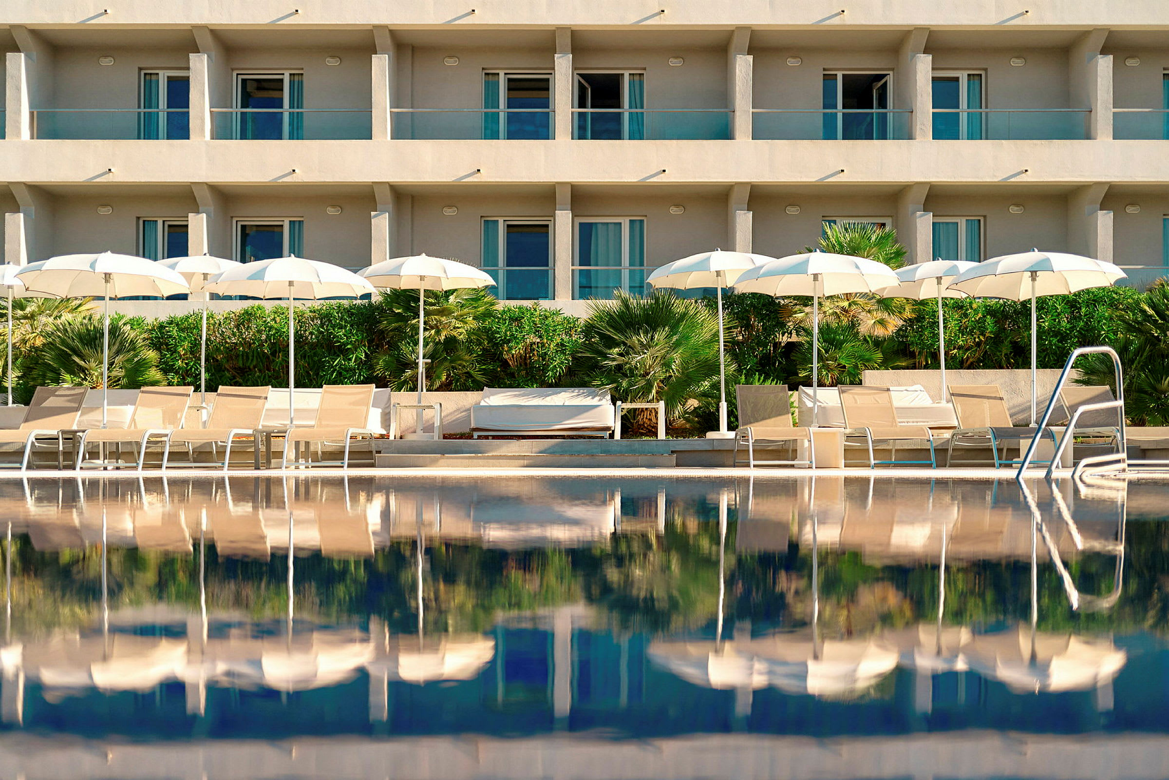 Bild på en upplyst hotellfasad med balkonger bakom en pool. Framför poolen finns solstolar och vita parasoller samt grönska. (AI-genererad text)