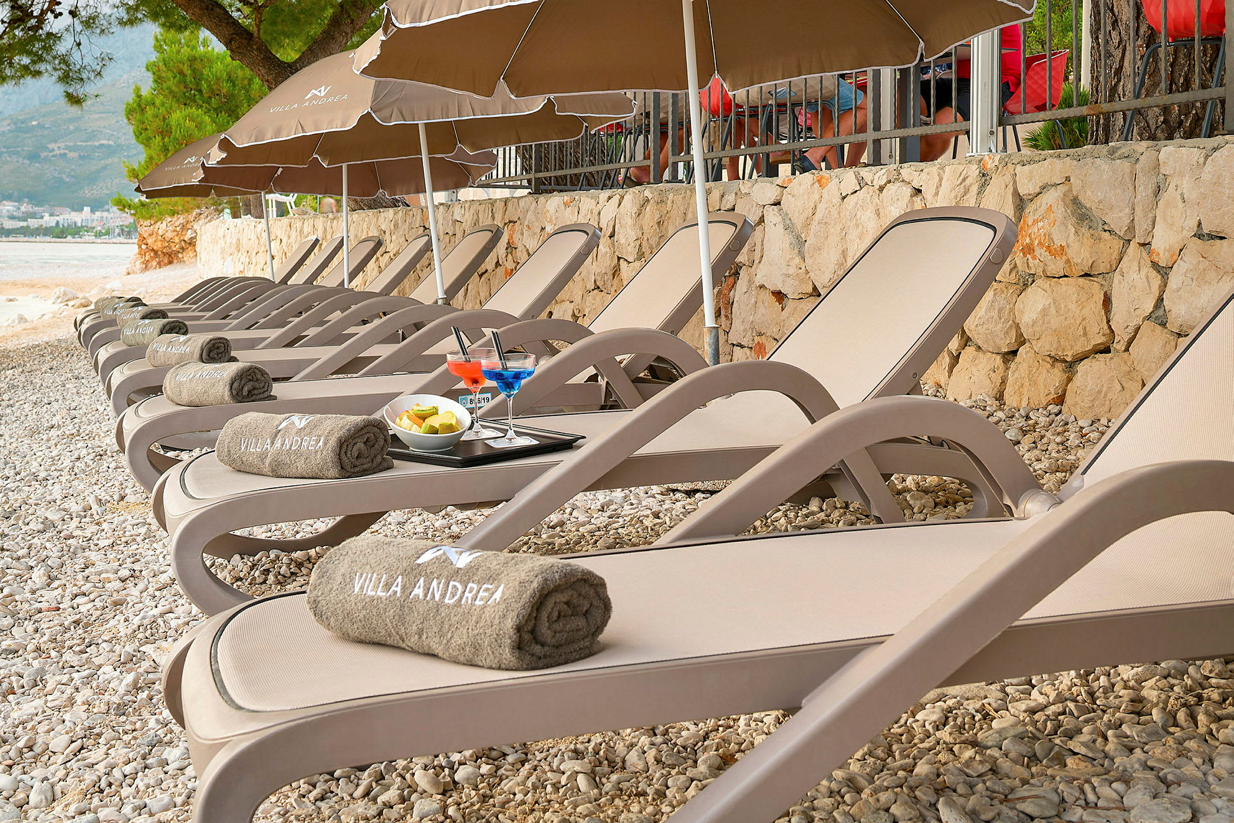 Rad av solsängar på en stenstrand, med handdukar märkta 'Villa Andrea', drinkar och frukt på en bricka. (AI-genererad text)