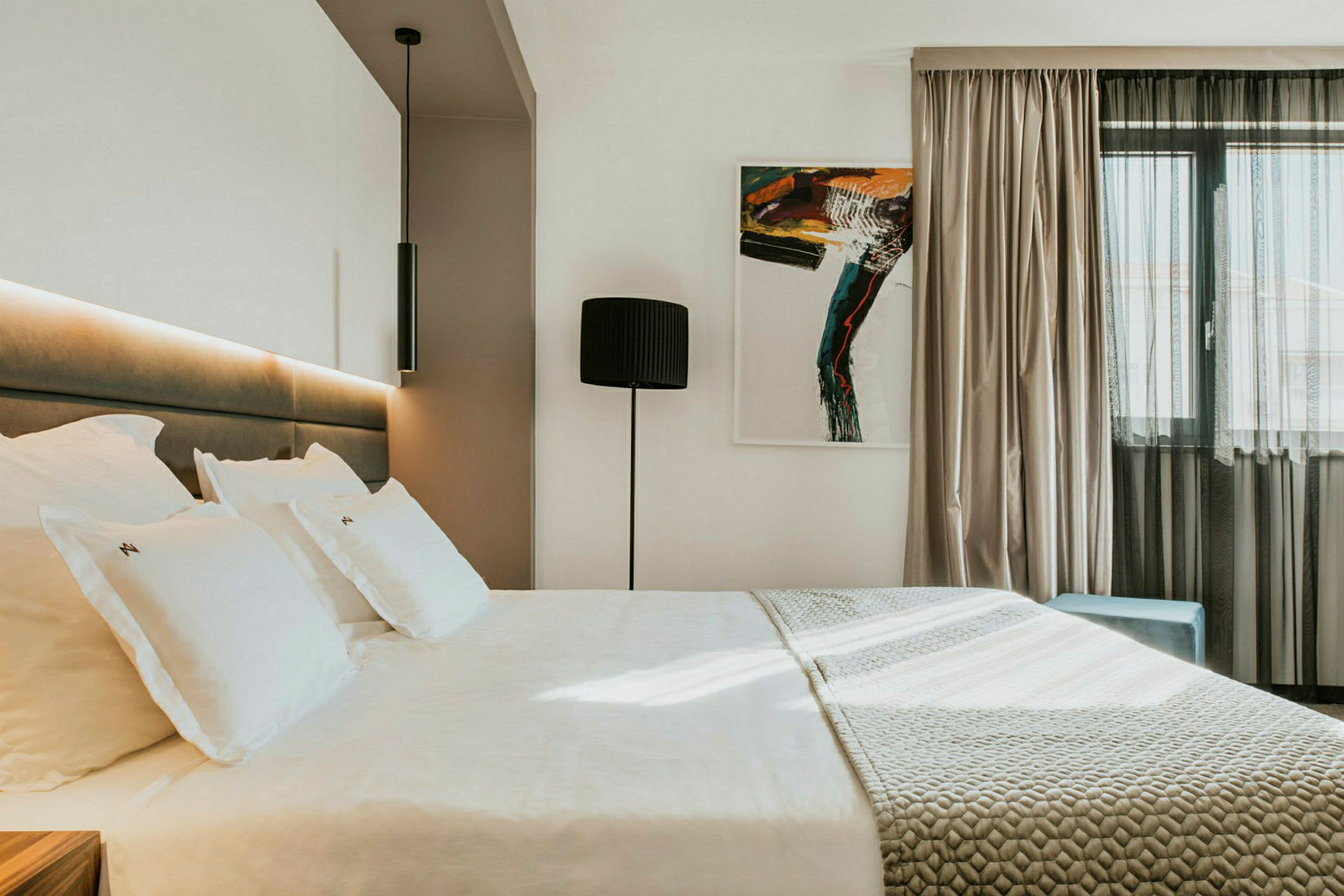 Modern hotellrum med en stor säng, konstverk på väggen och en fönster med ljusa gardiner. (AI-genererad text)