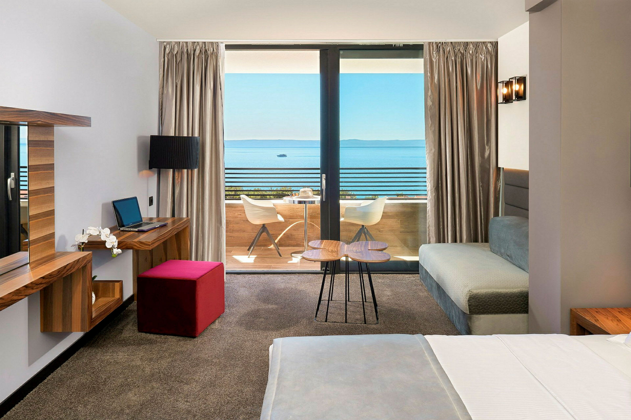 Hotellrum med säng, skrivbord och utsikt mot en balkong och hav. (AI-genererad text)