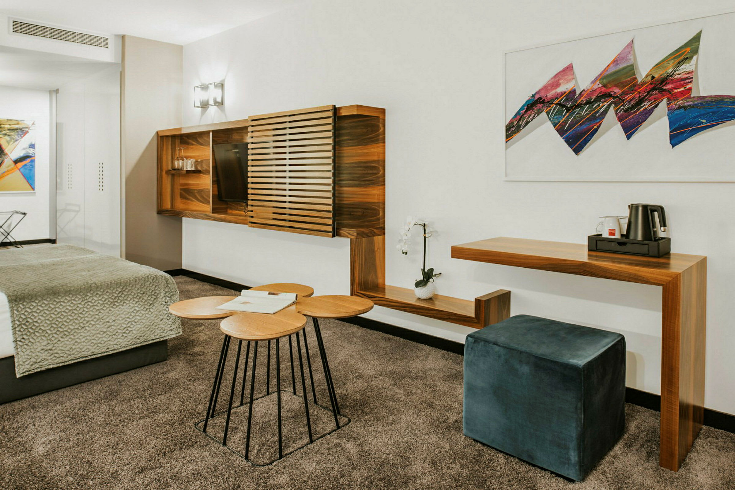 Ett modernt hotellrum med möbler i trä, en sittpuff, och konst på väggen. (AI-genererad text)