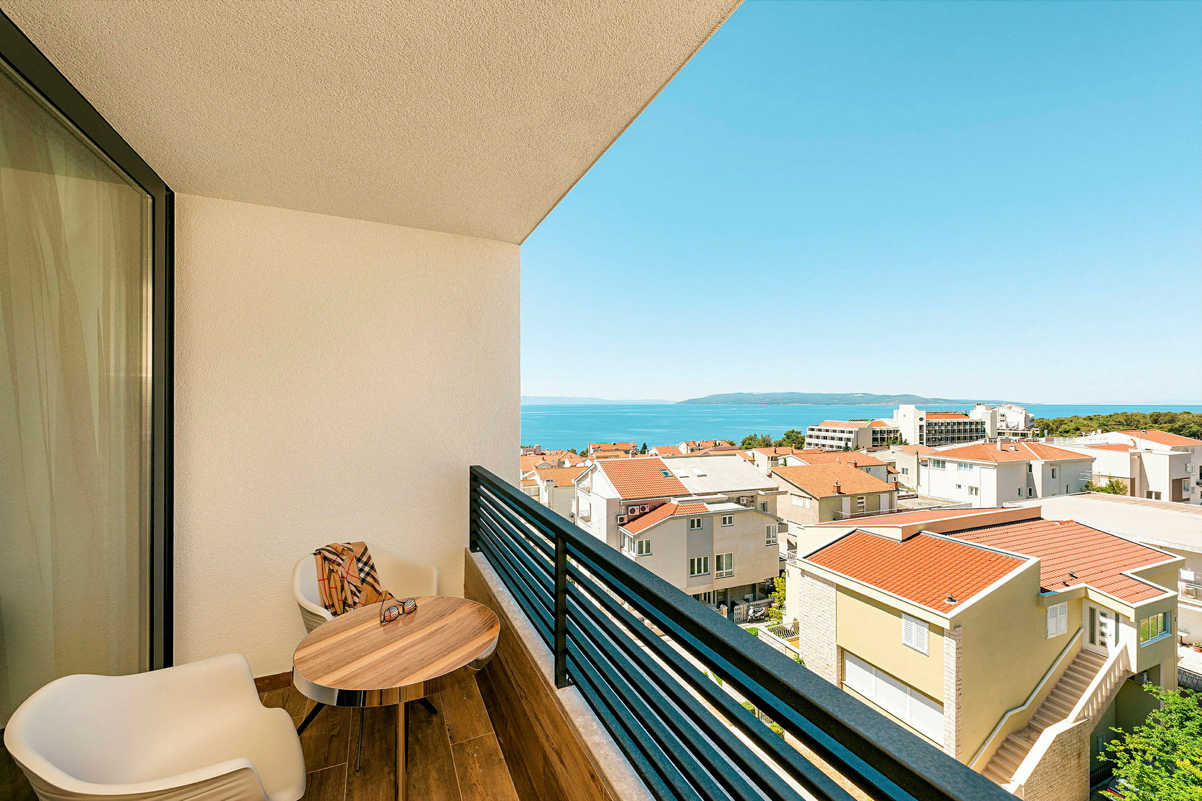 Utsikt från en balkong med två stolar, ett bord och utsikt över en stadsbebyggelse och havet. (AI-genererad text)
